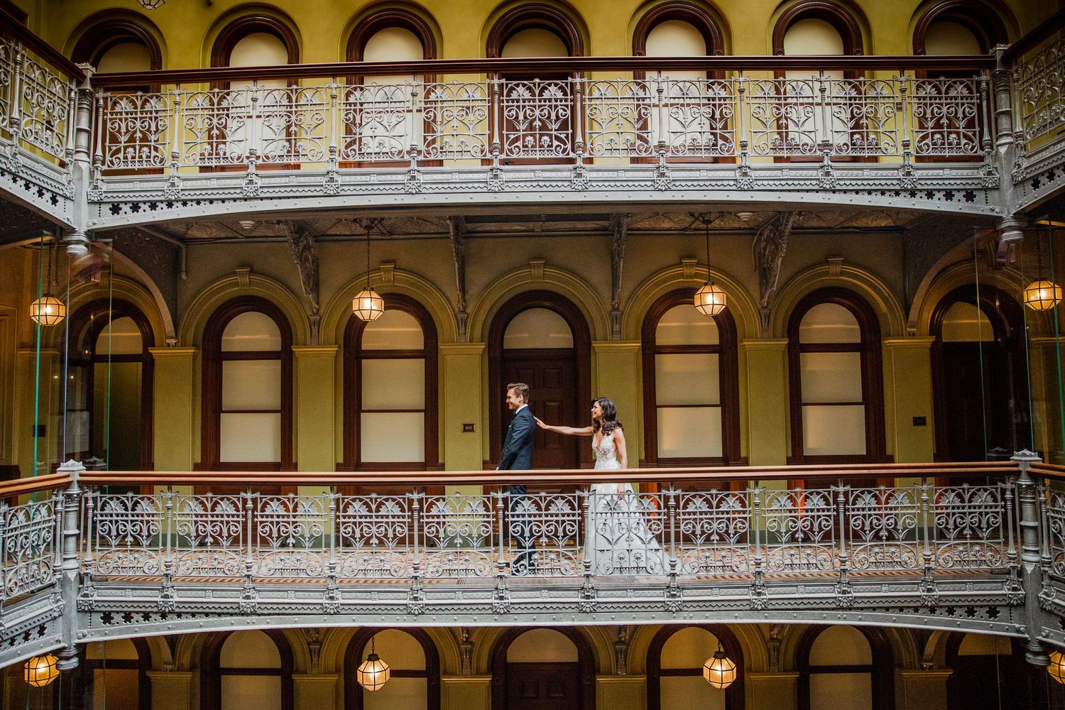 An elegant wedding couple stands on an ornate Victorian balcony with arched windows and decorative railings.