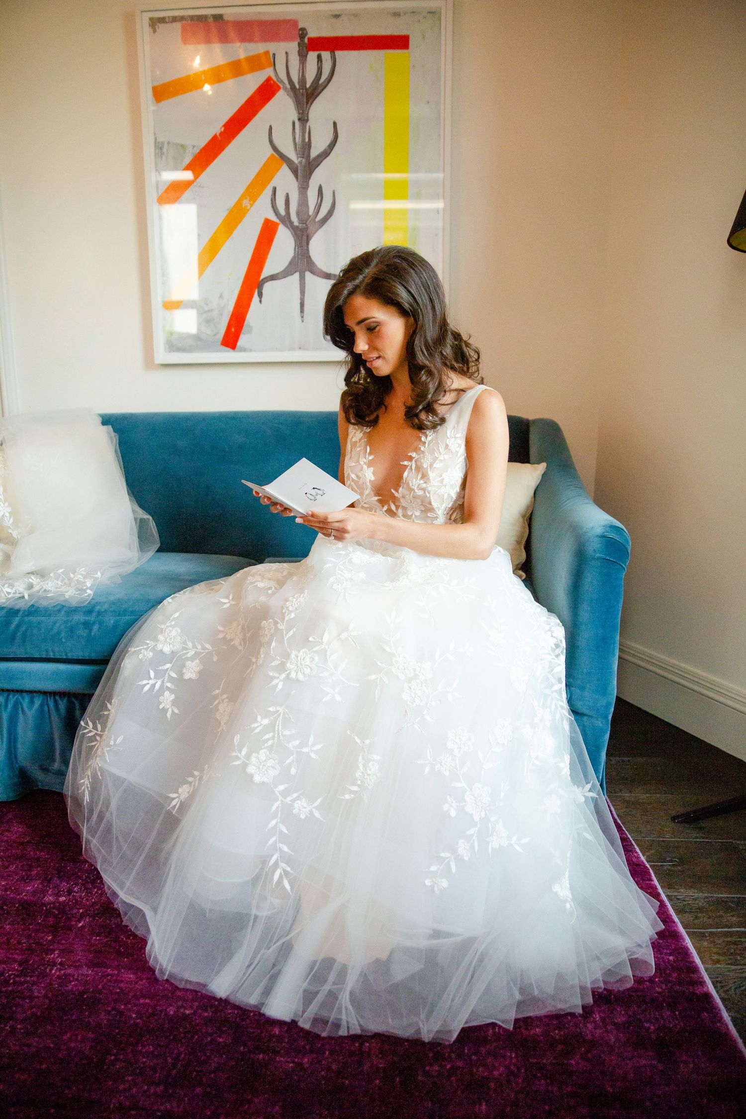 Bride in lace gown reading letter while seated on blue velvet couch.