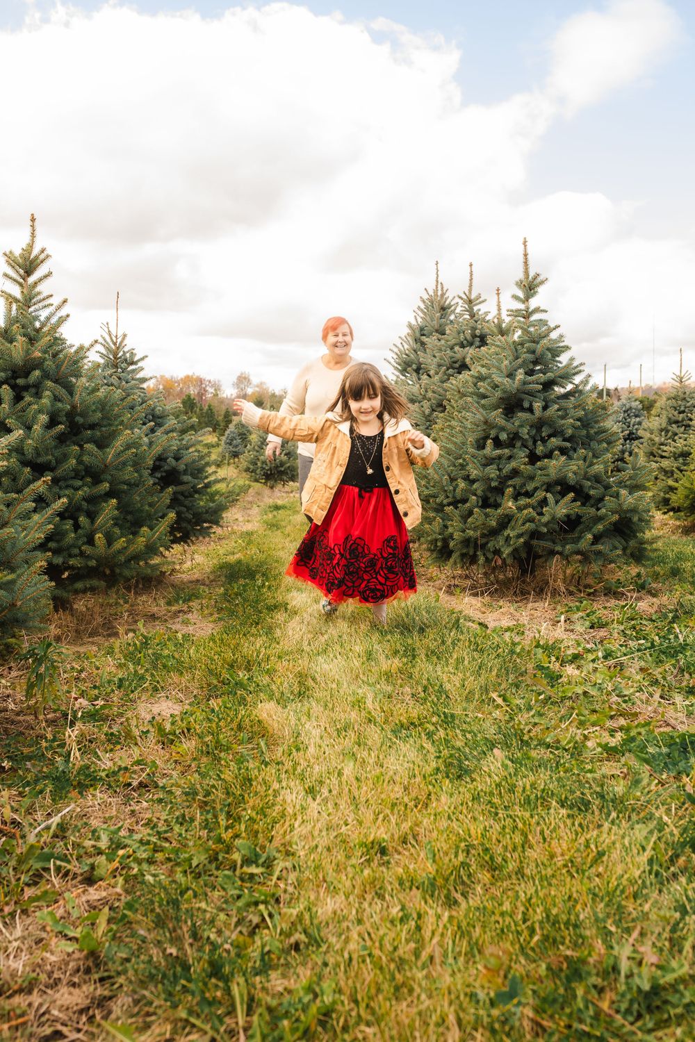 A small child wearing a red dress runs through a grassy field at a Christmas tree farm on a cloudy day.