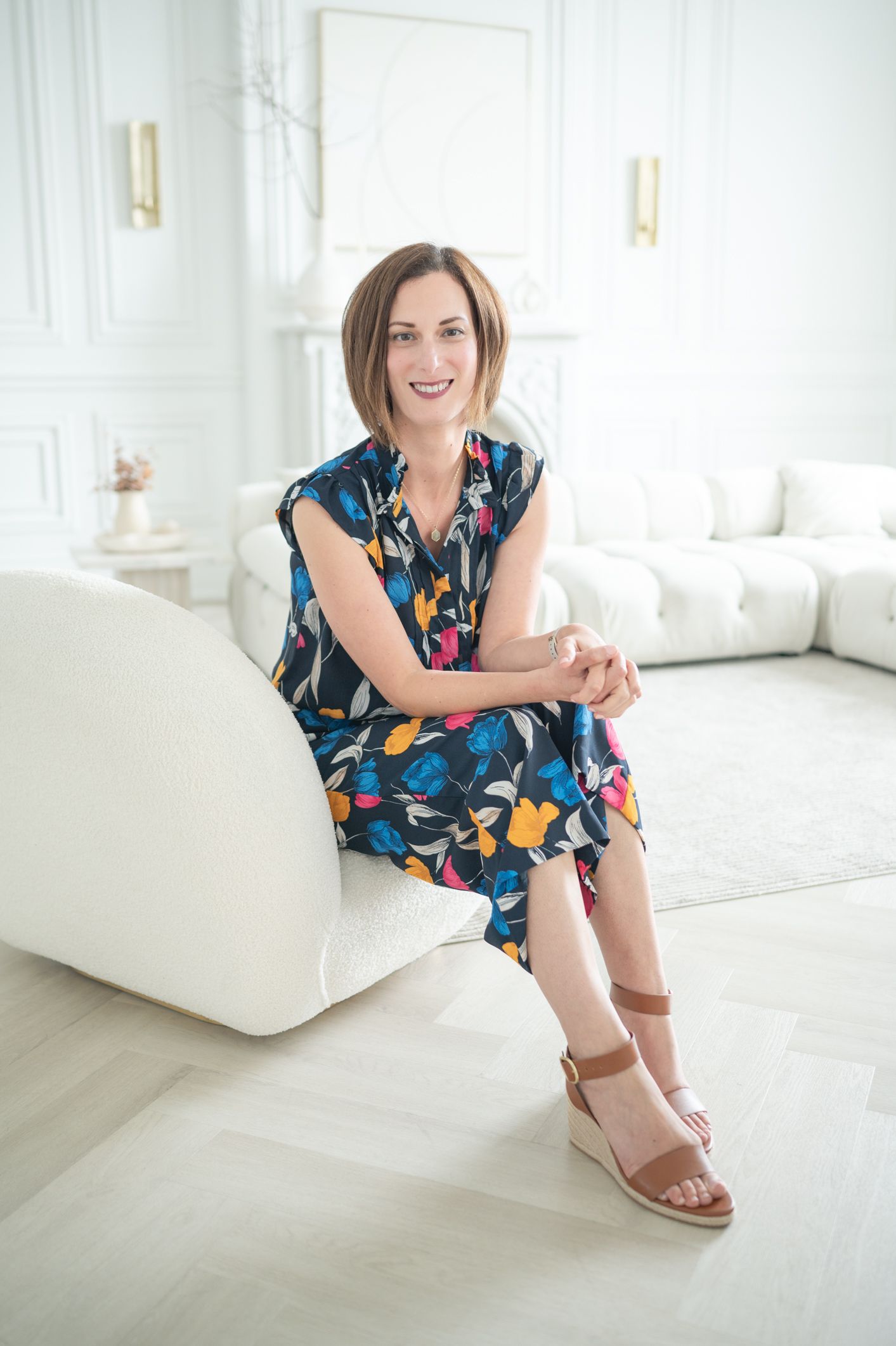 Woman in colorful floral dress sitting on white couch in bright modern room.