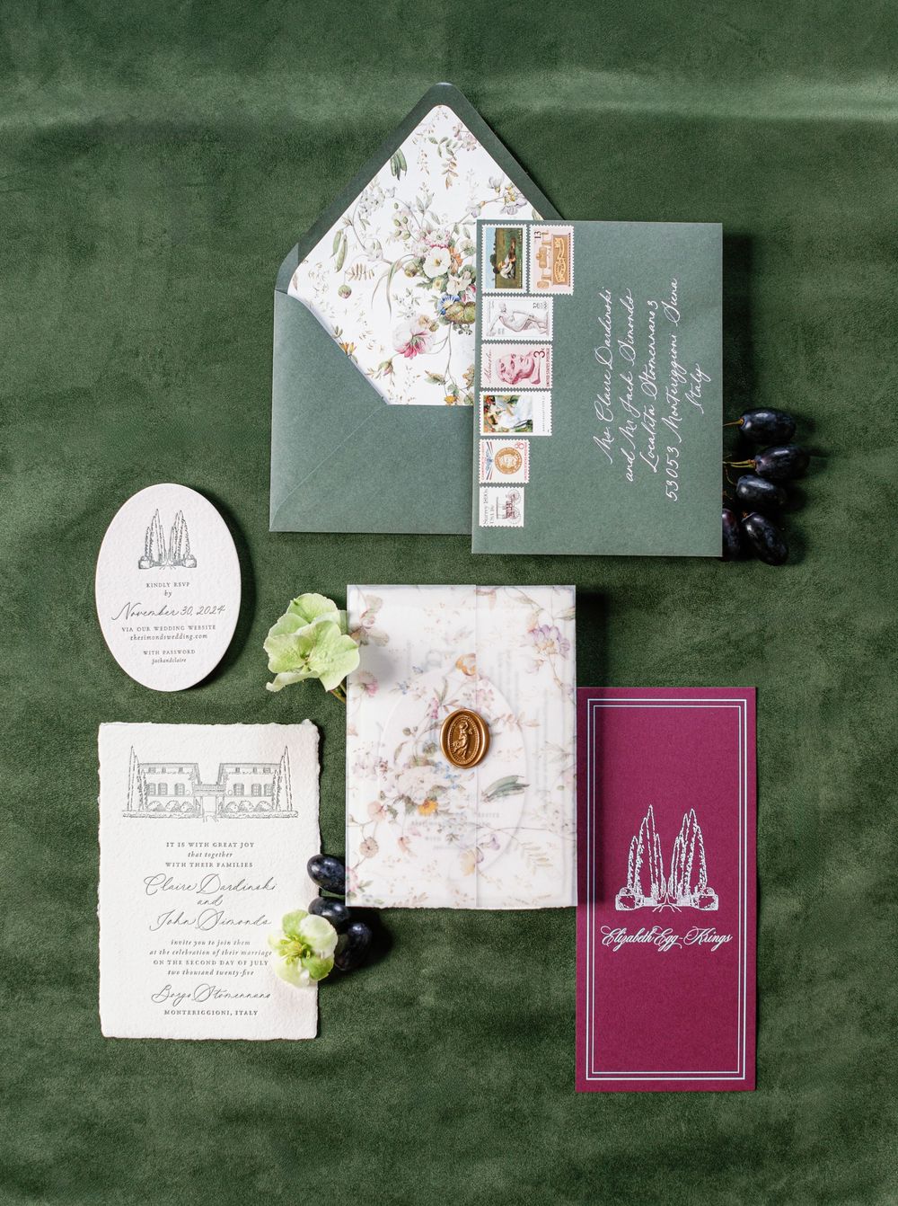 Wedding stationery suite with floral designs and maroon velvet books arranged elegantly on dark green velvet background.