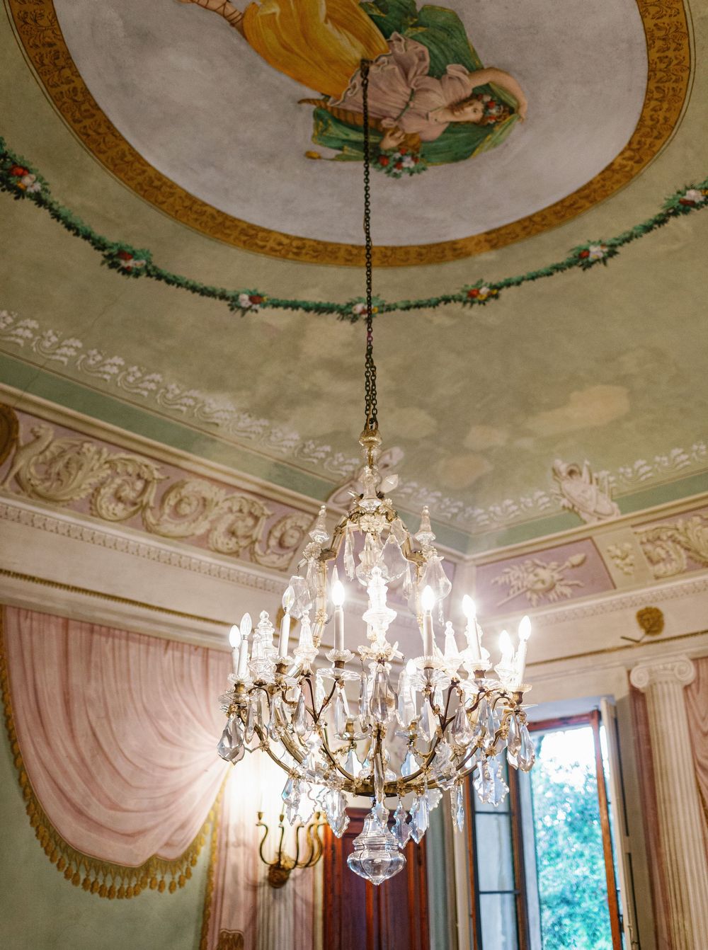 Crystal chandeliers hang from ornately decorated ceiling medallions in a historic grand room with gold trim details.