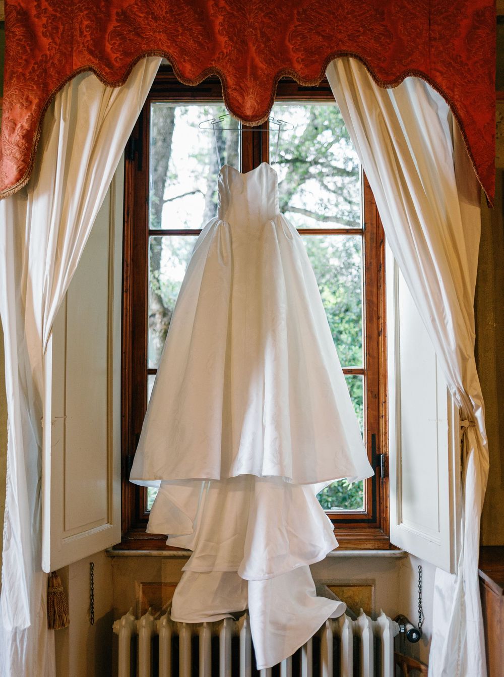 A white wedding dress Pnina Tornai hangs gracefully in front of a window with cream curtains and a red valance, next to an antique radiator.