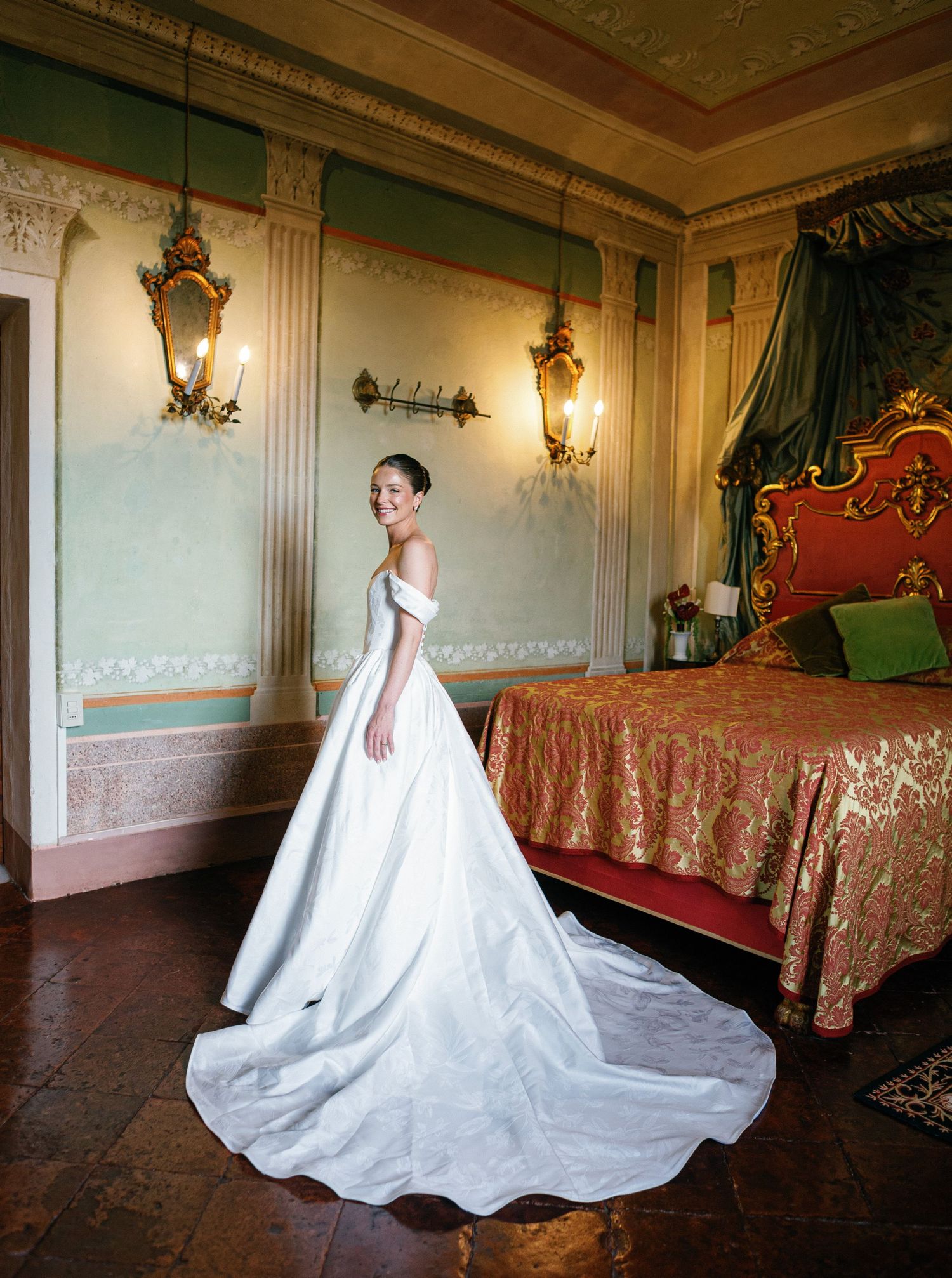A bride in a flowing white wedding gown with dramatic train poses elegantly in an ornate historic interior ballroom.