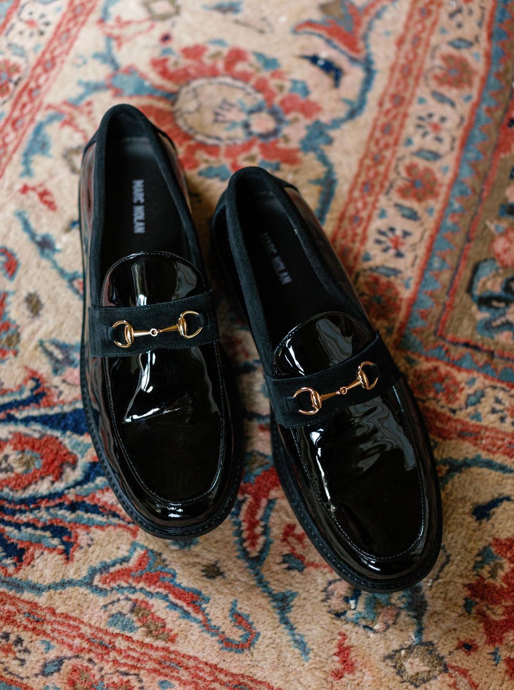 A pair of black patent leather loafers with gold horsebit hardware on an ornate oriental rug.