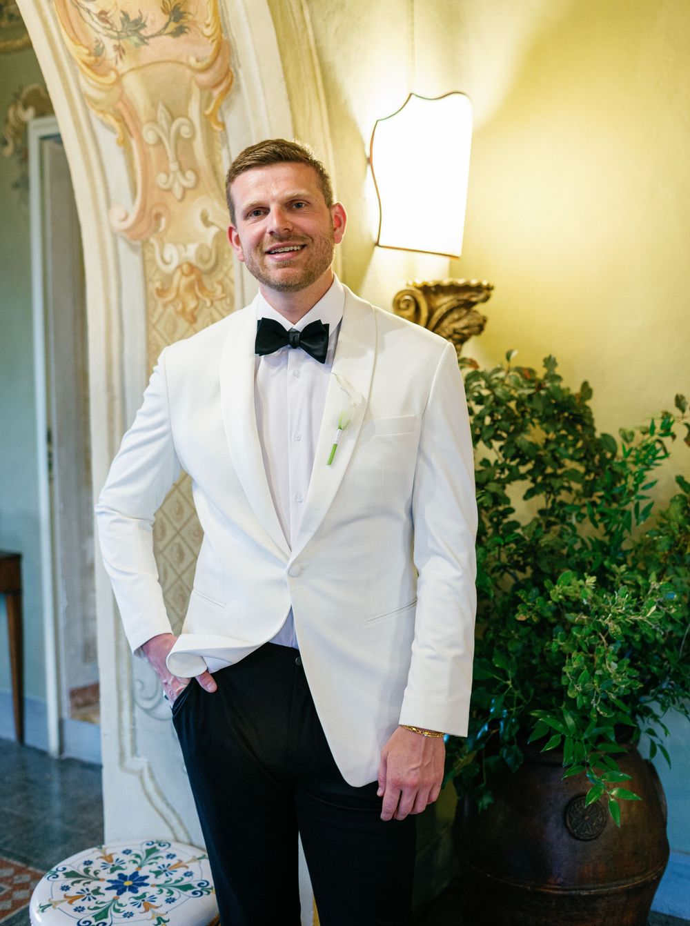 A groom in a white dinner jacket and black bow tie poses in an elegant hallway with ornate walls and greenery.