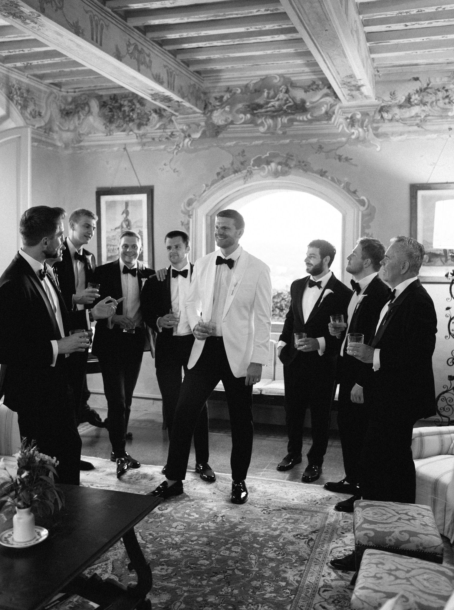A panoramic black and white photo shows a group of men in formal tuxedos gathering in an elegant room.