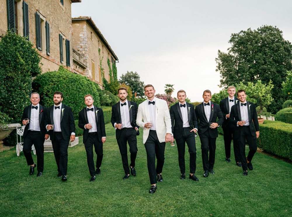 Group of groomsmen in black tuxedos walk across a lush green lawn outside a stone building.