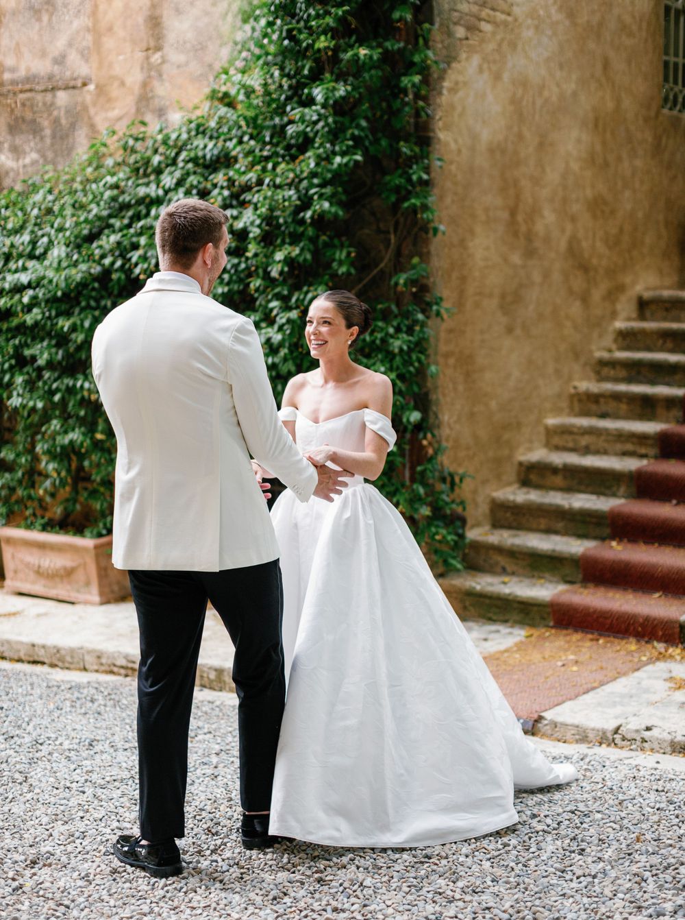 Two people in formal wedding attire sharing a tender moment in front of a rustic stone wall and staircase.