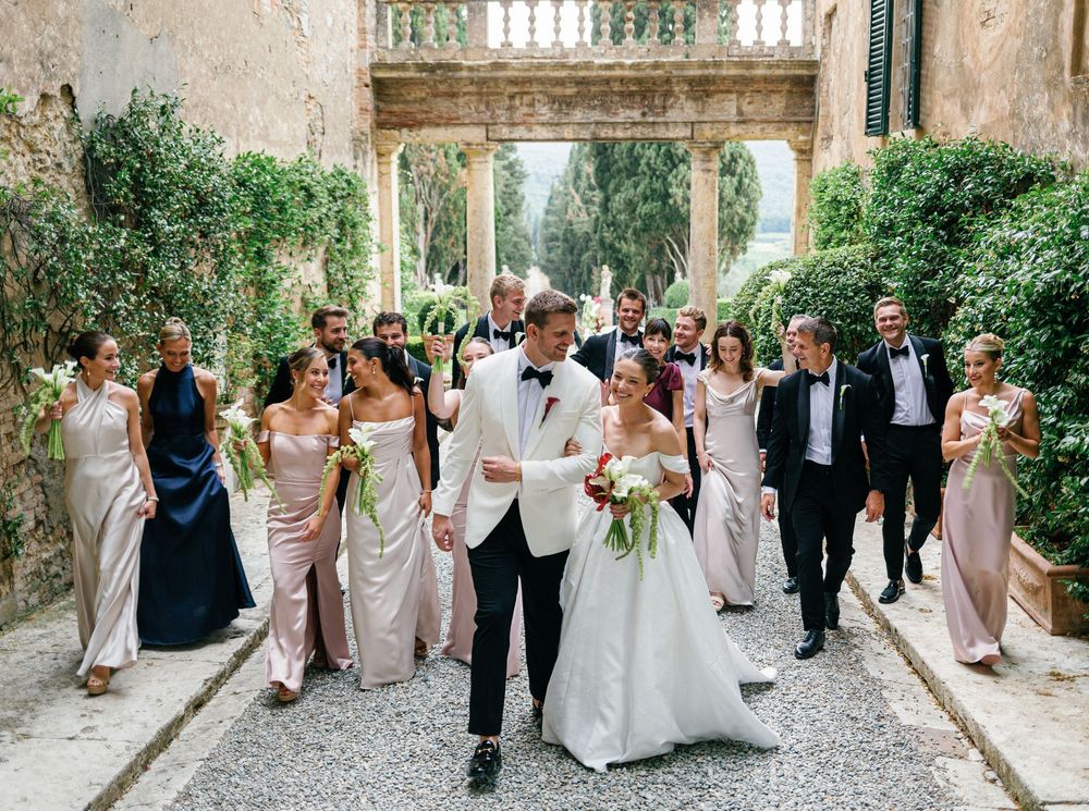 Wedding party walking and laughing together along a stone path bordered by elegant archways and lush greenery.