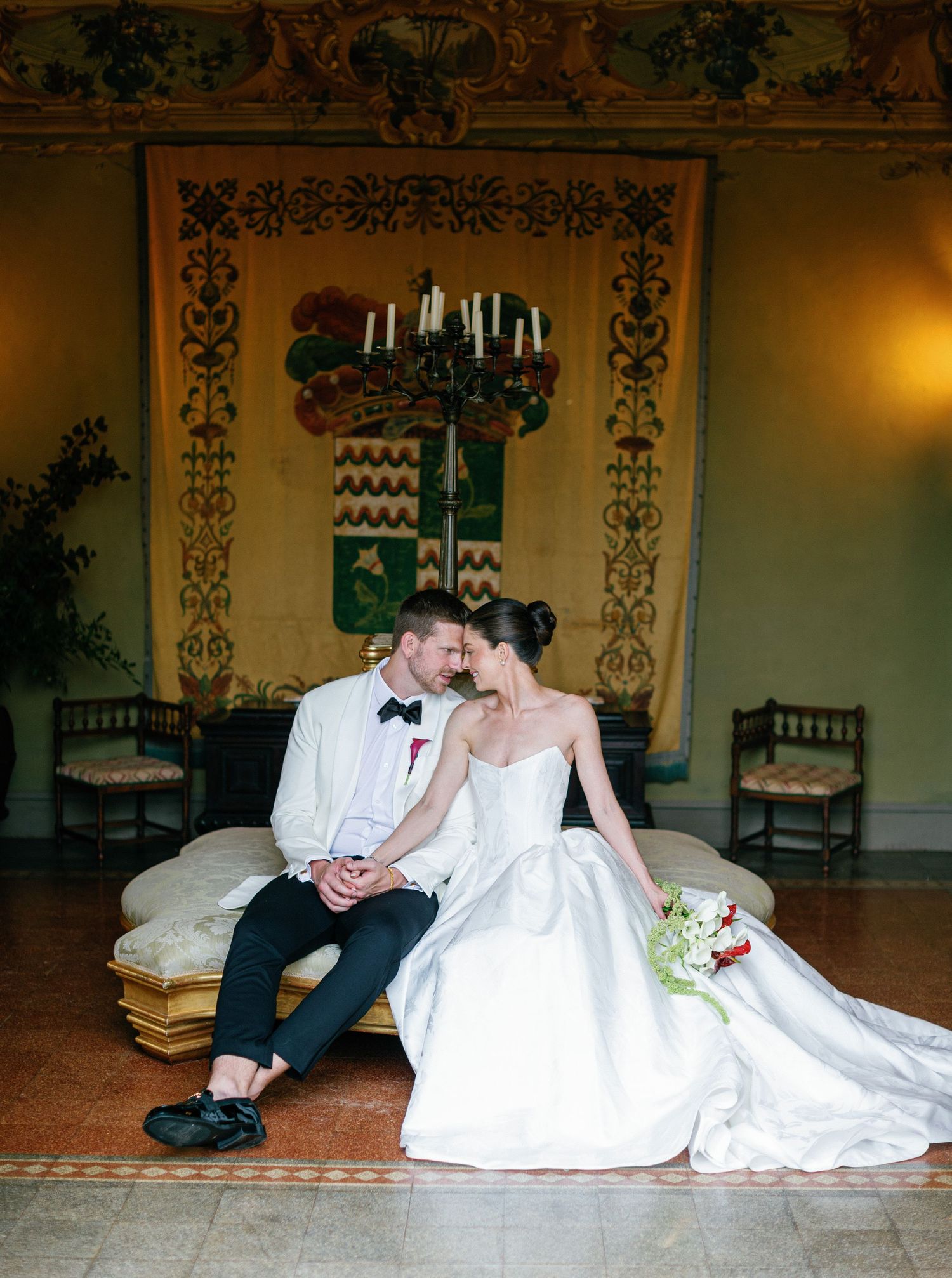 A couple in traditional wedding attire sits close together in an elegant room with ornate wall decorations and a chandelier.