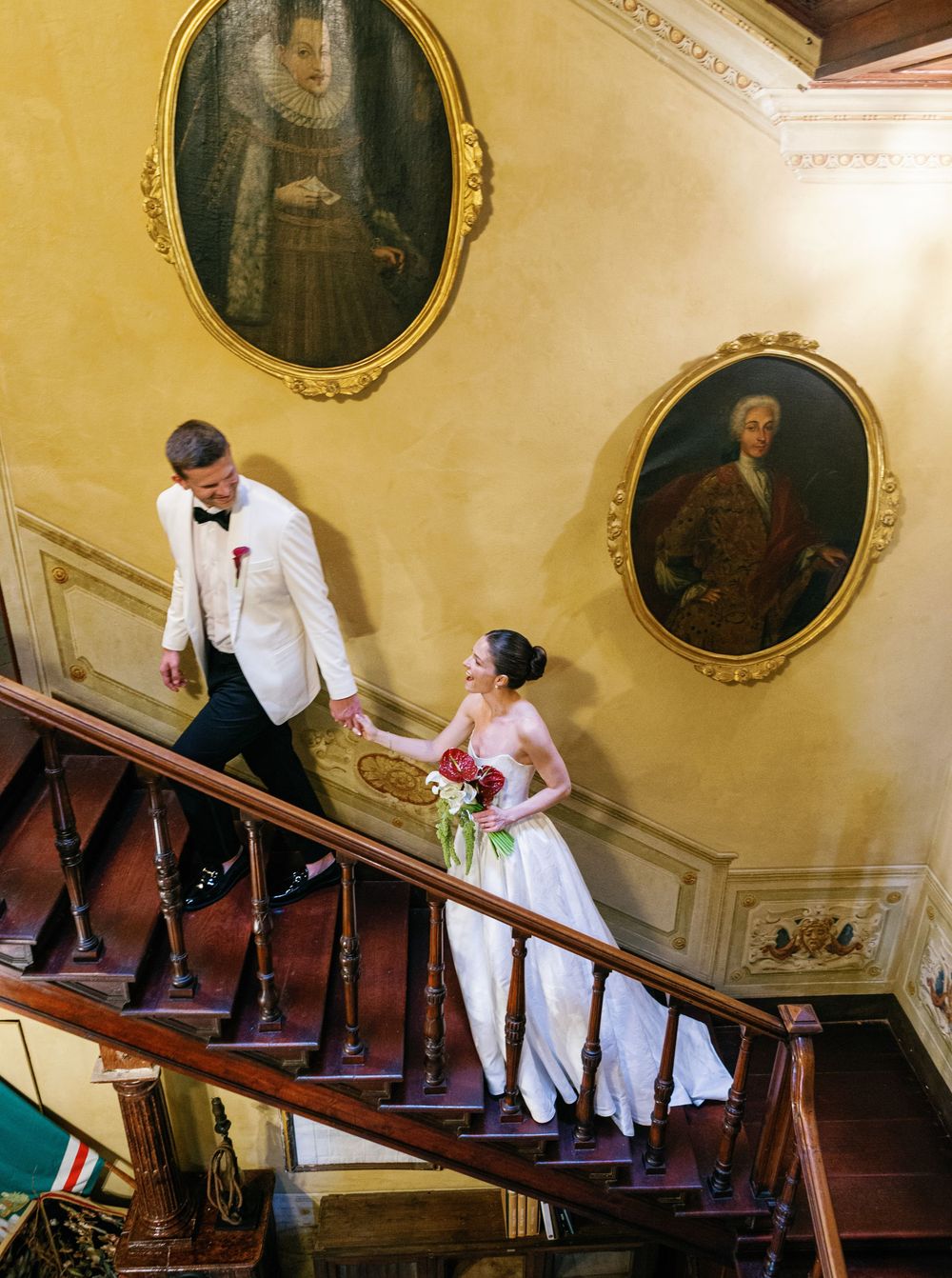 Wedding couple descends elegant staircase with historic portraits on wall in mansion setting.