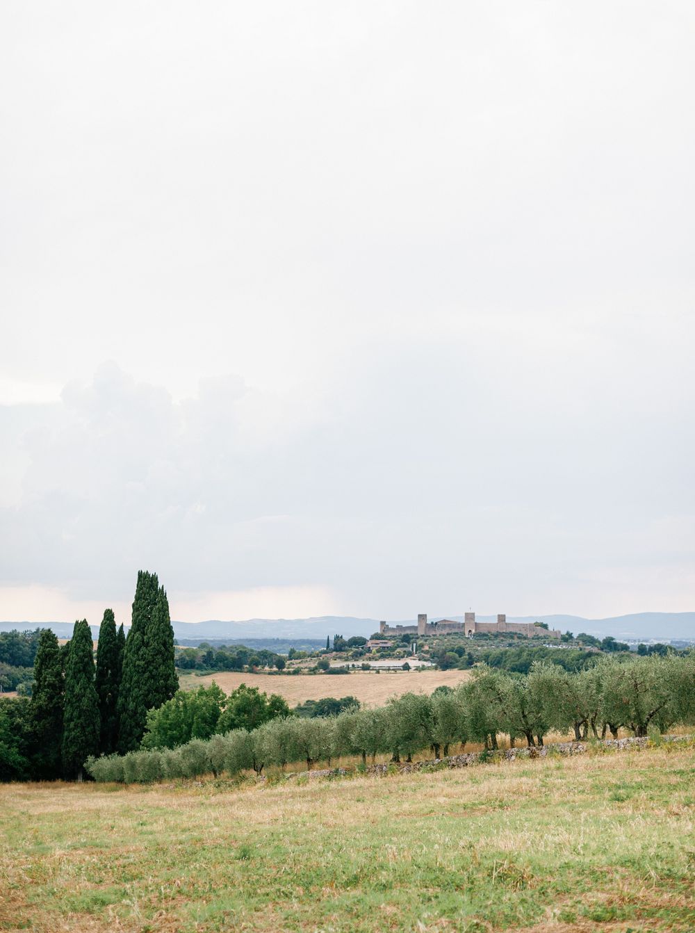 Panoramic view of rolling Tuscan hills with cypress trees and distant countryside buildings under cloudy skies.