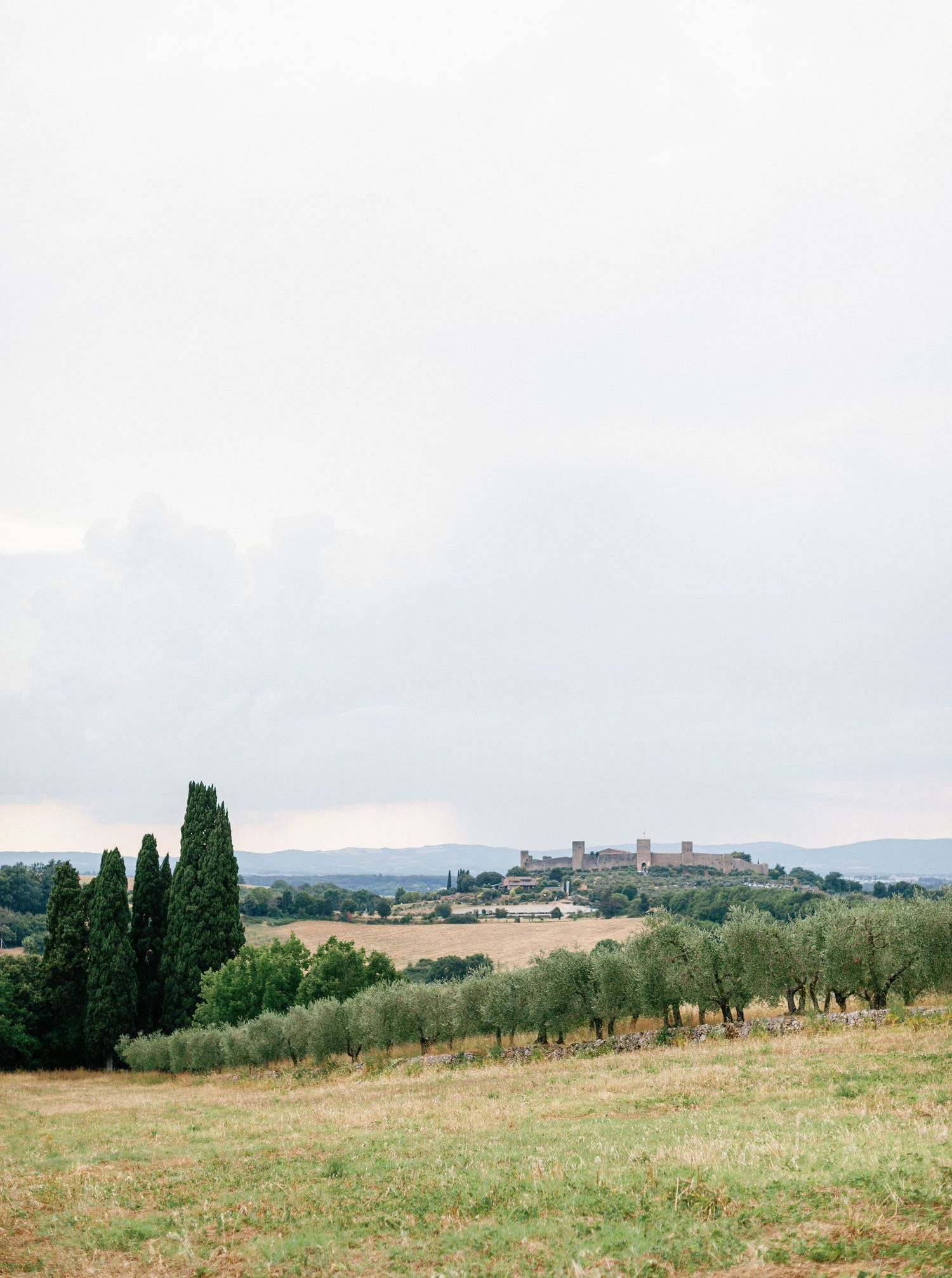 Panoramic view of rolling Tuscan hills with cypress trees and distant countryside buildings under cloudy skies.