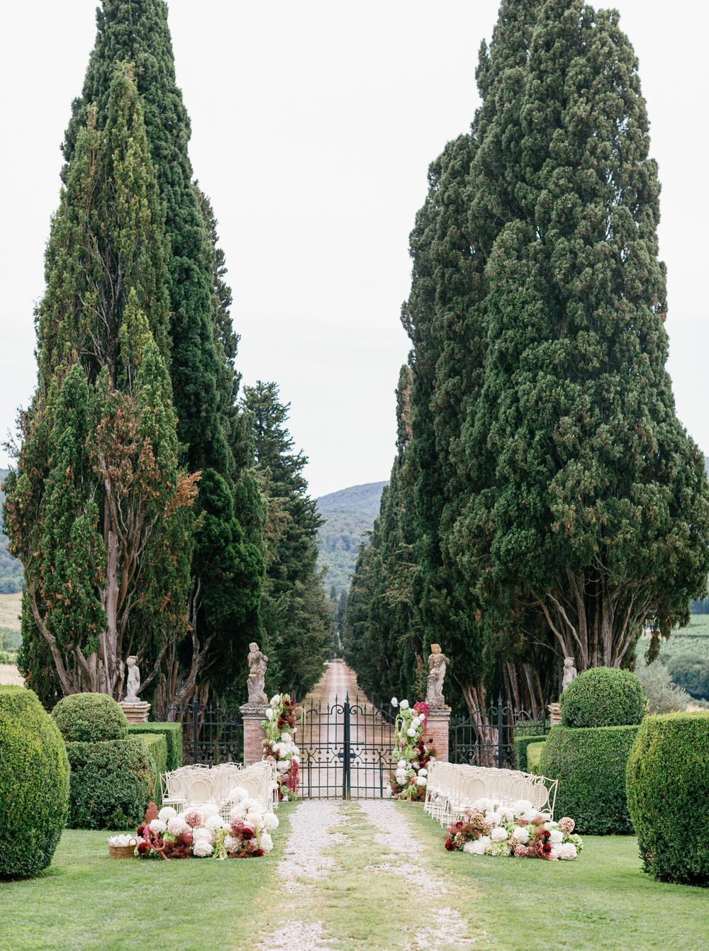 Wedding ceremony at Borgo Stomennano in garden path lined with tall cypress trees and manicured hedges leads to a distant mountain vista.