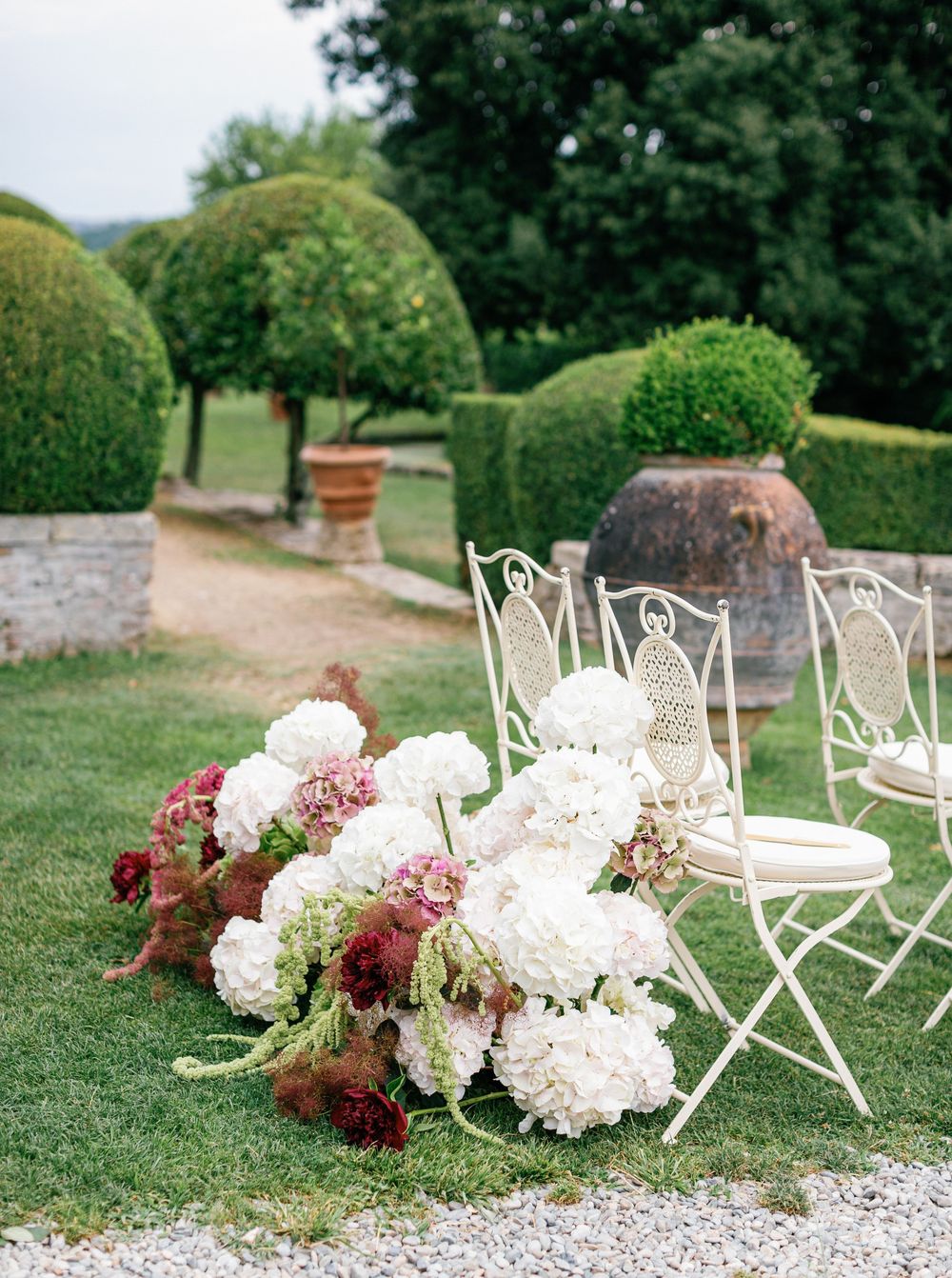 White garden chairs and elegant floral arrangements with pink and burgundy blooms line a gravel pathway in an outdoor setting.