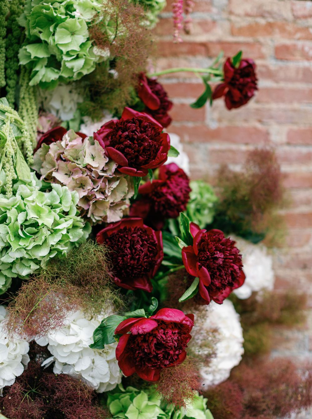 A stunning floral arrangement featuring deep red carnations, white hydrangeas, and green blooms against a rustic brick wall.