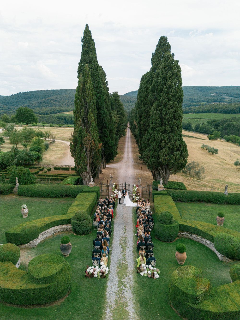 A cypress-lined pathway through Tuscan countryside at Borgo Stomennano with guests gathered in the garden, captured in multiple drone views.