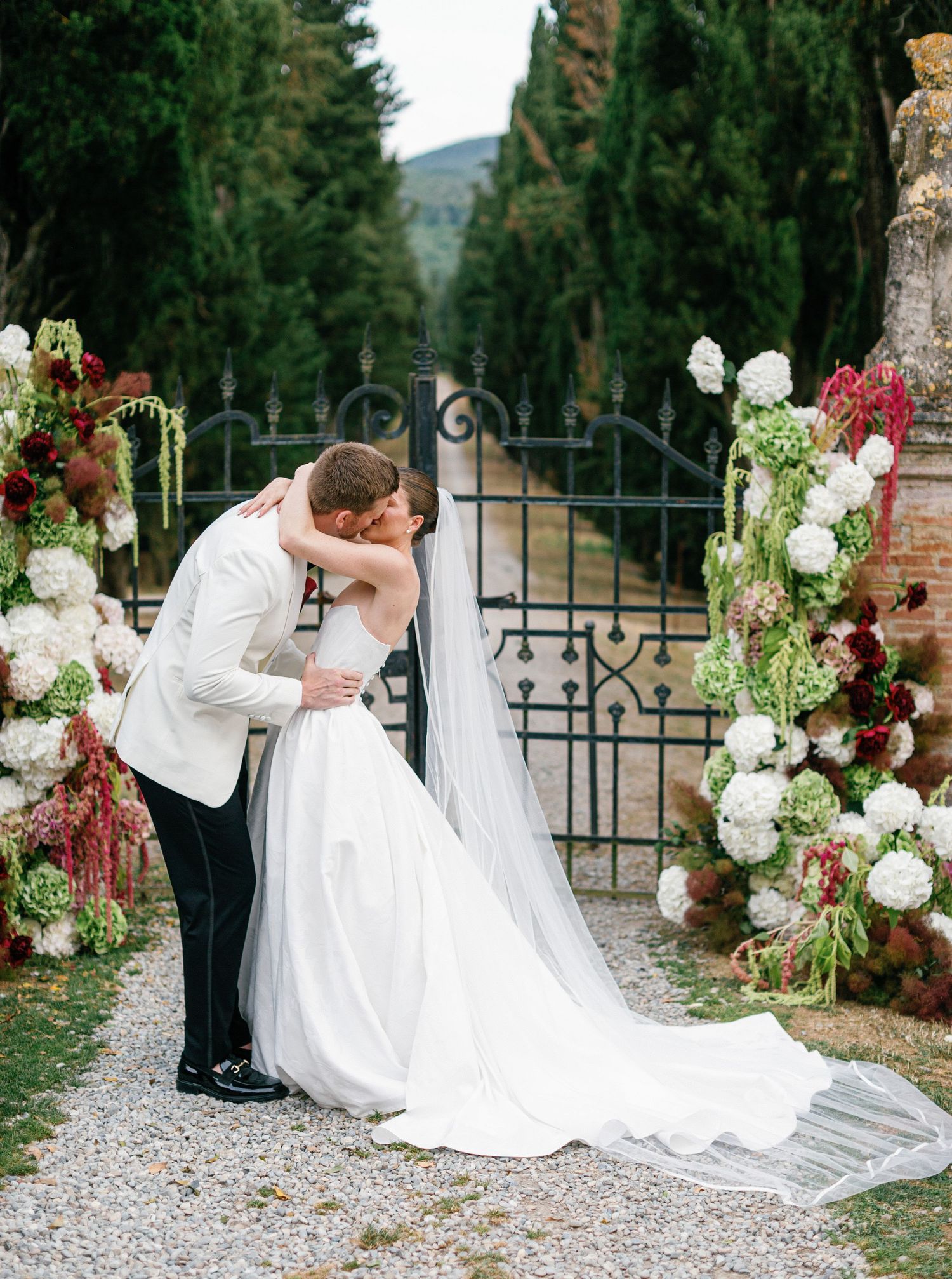A romantic wedding moment captured in a garden setting with wrought iron gates and floral decorations in burgundy and white.