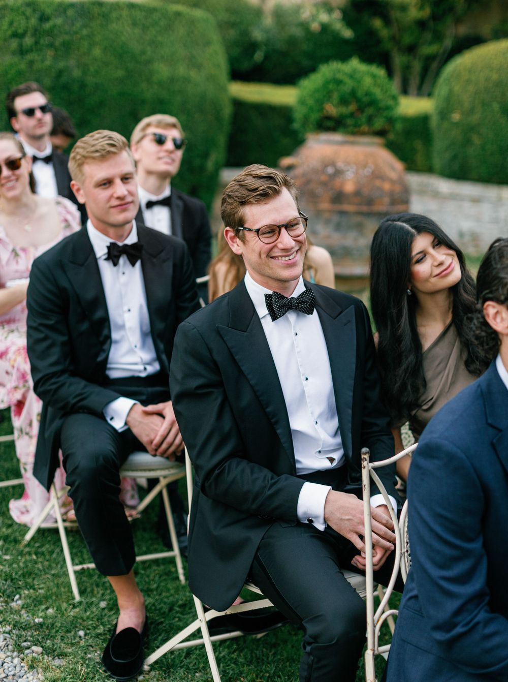 Wedding guests in formal black tie attire sit on white wooden chairs during an outdoor garden ceremony.