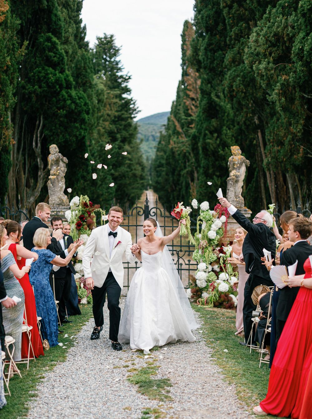 Newlyweds walk down a cypress-lined path with guests celebrating at an elegant Italian villa Borgo Stomennano wedding ceremony.