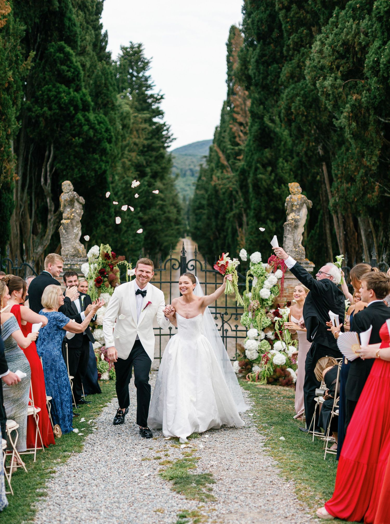 Newlyweds walk down a cypress-lined path with guests celebrating at an elegant Italian villa Borgo Stomennano wedding ceremony.