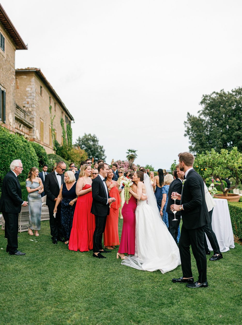 Wedding guests gather in a semicircle formation on a green lawn outside a rustic stone building during an outdoor ceremony.