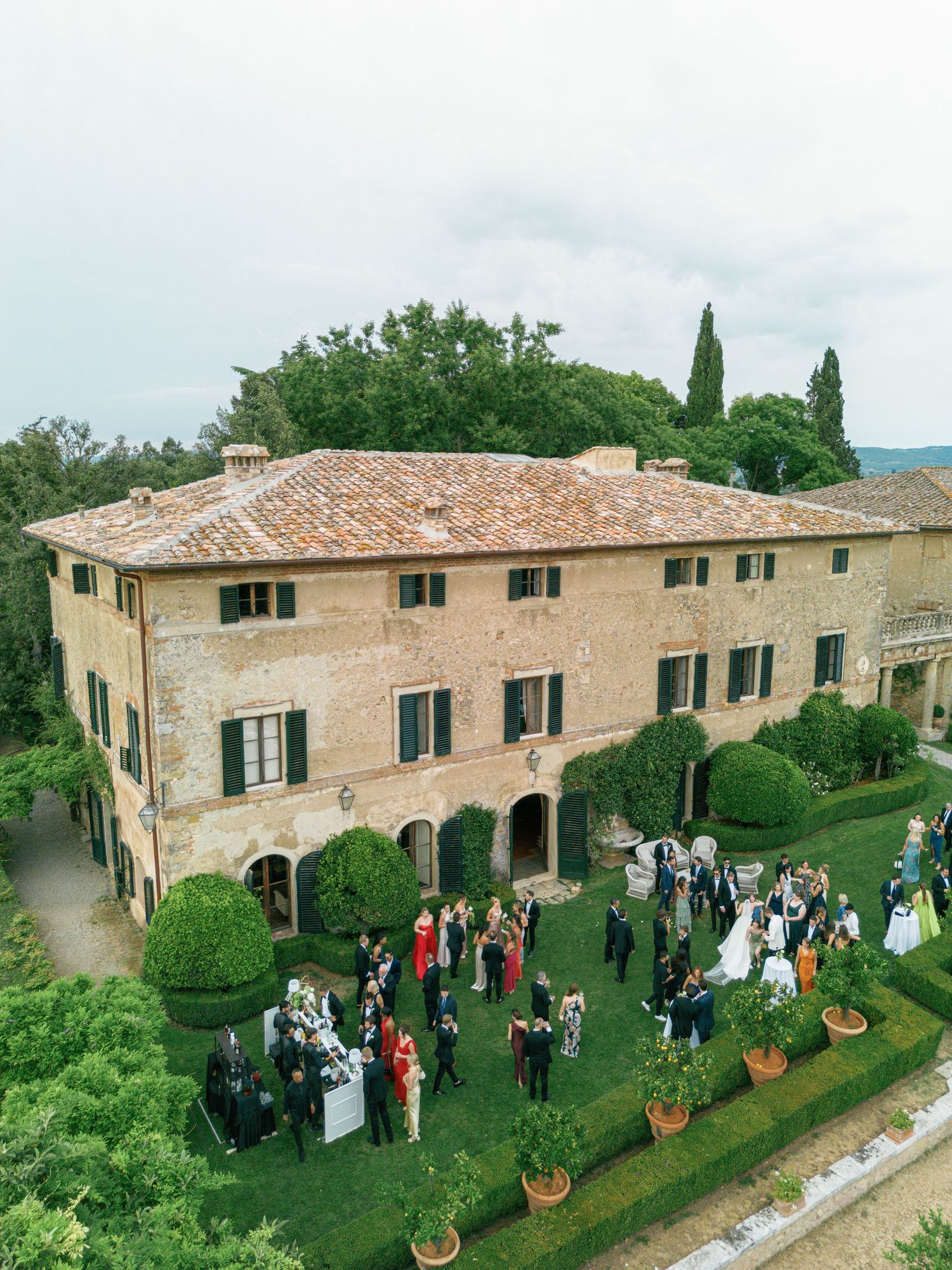 A panoramic view of a rustic Tuscan villa Borgo Stomennano with terra cotta roof hosting an outdoor gathering on its manicured lawn.
