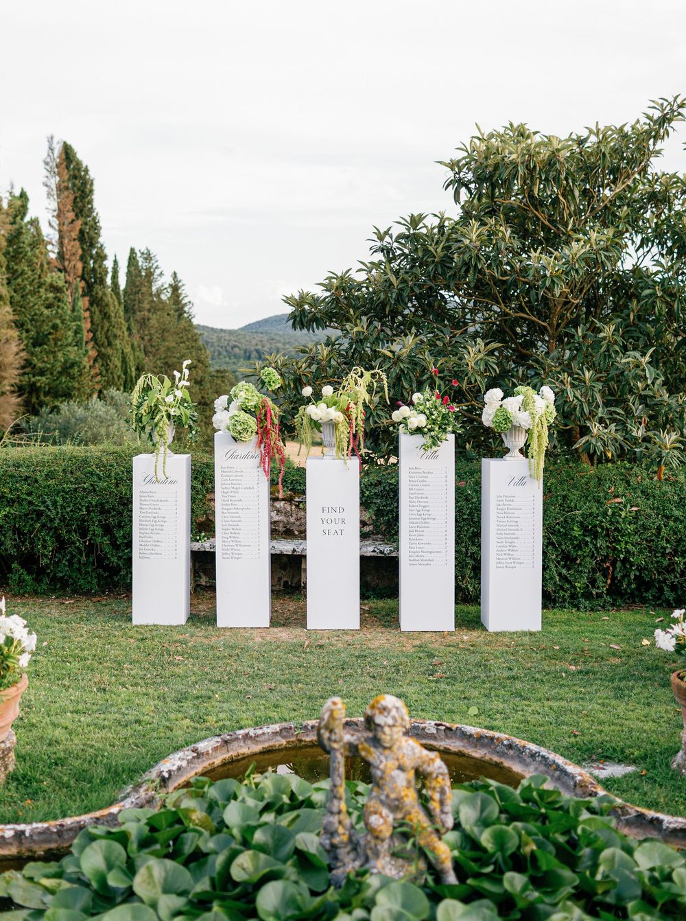 A row of elegant white seating charts displayed on grass with greenery and trees in the background.
