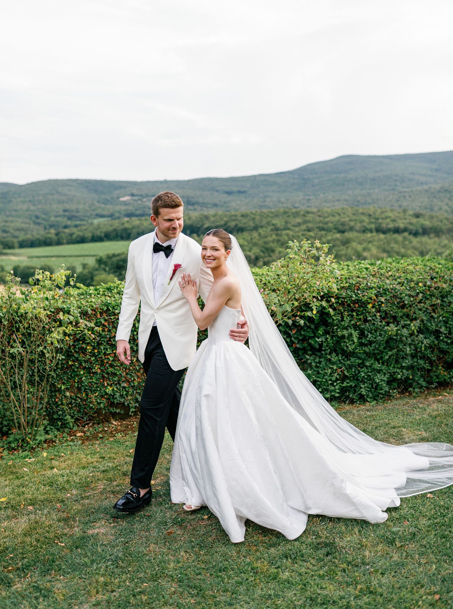 Couple in wedding attire walking together on hillside path with flowing white dress and veil in beautiful mountain scenery.