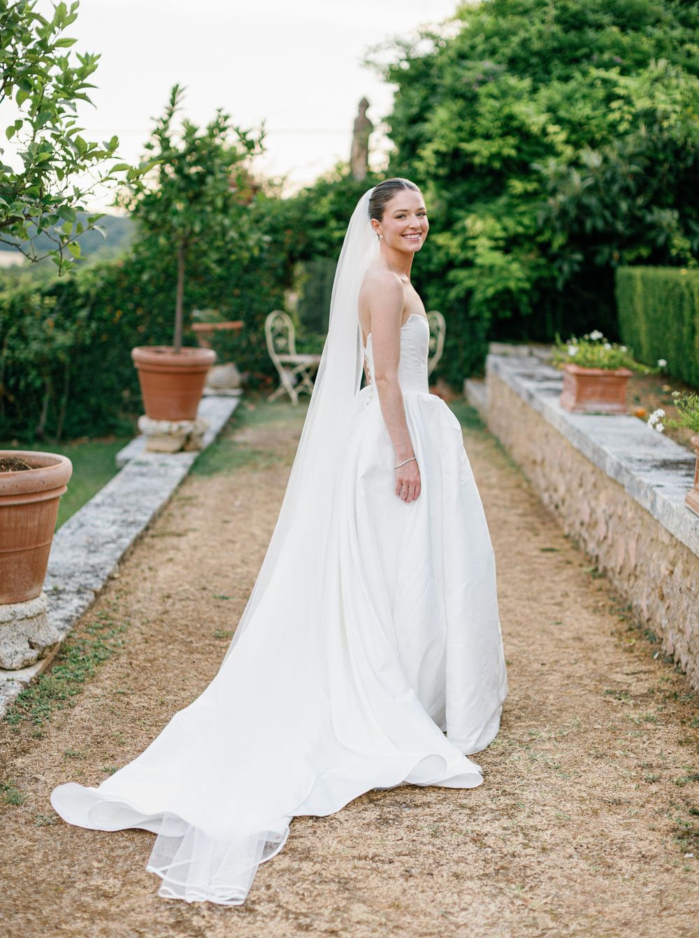 A flowing white wedding dress with lace details trails elegantly along a rustic garden pathway bordered by stone walls.