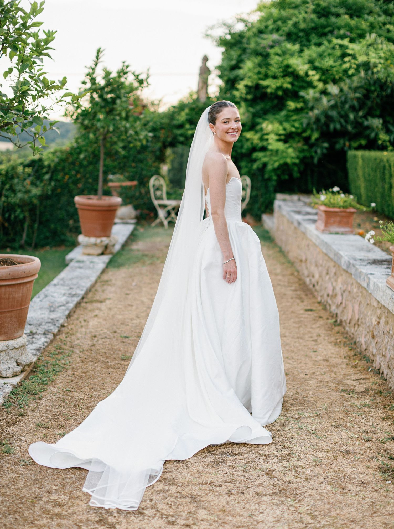 A flowing white wedding dress with lace details trails elegantly along a rustic garden pathway bordered by stone walls.