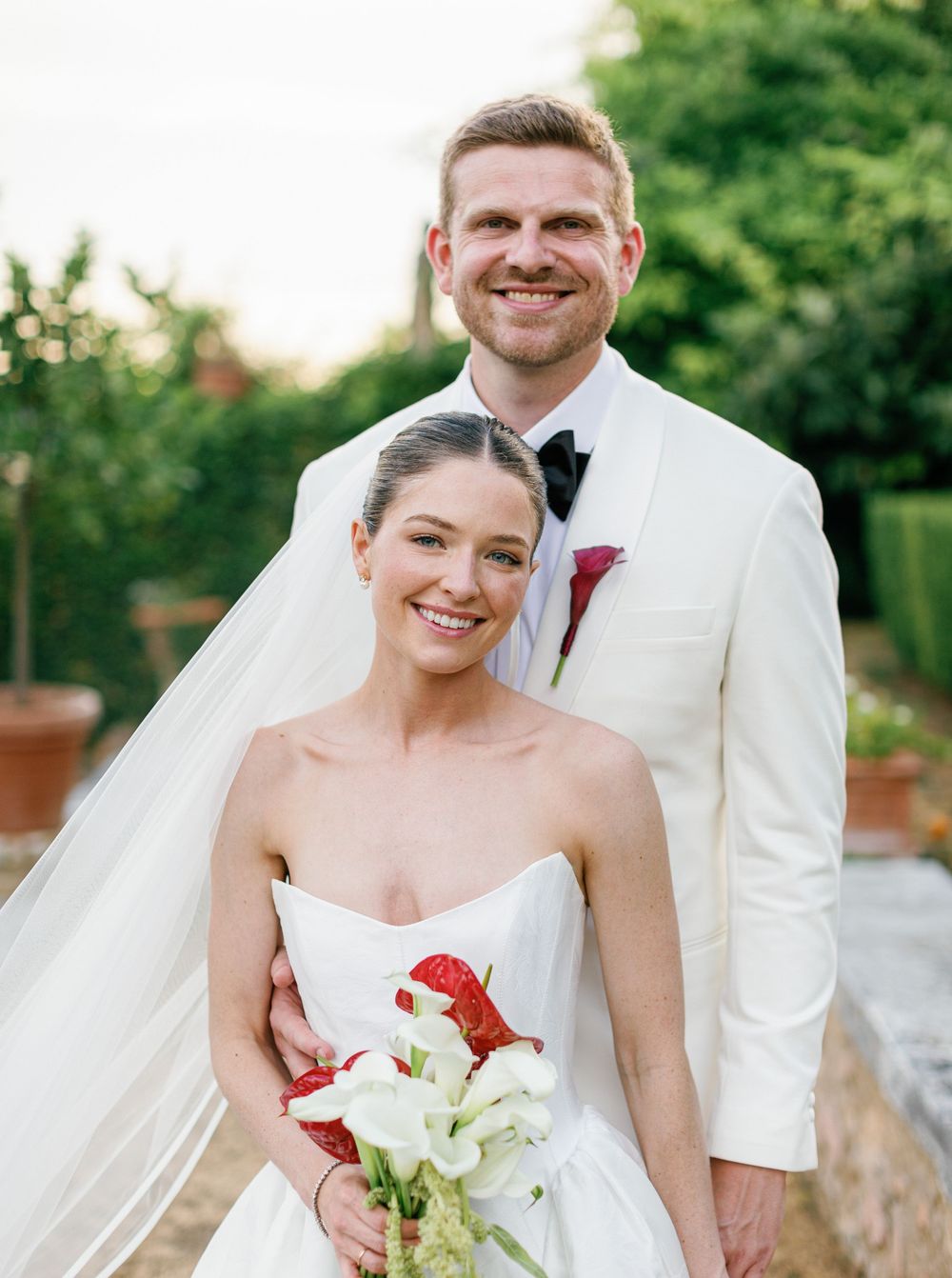 A wedding couple stands together outdoors with the bride holding a white lily bouquet and wearing a strapless gown.
