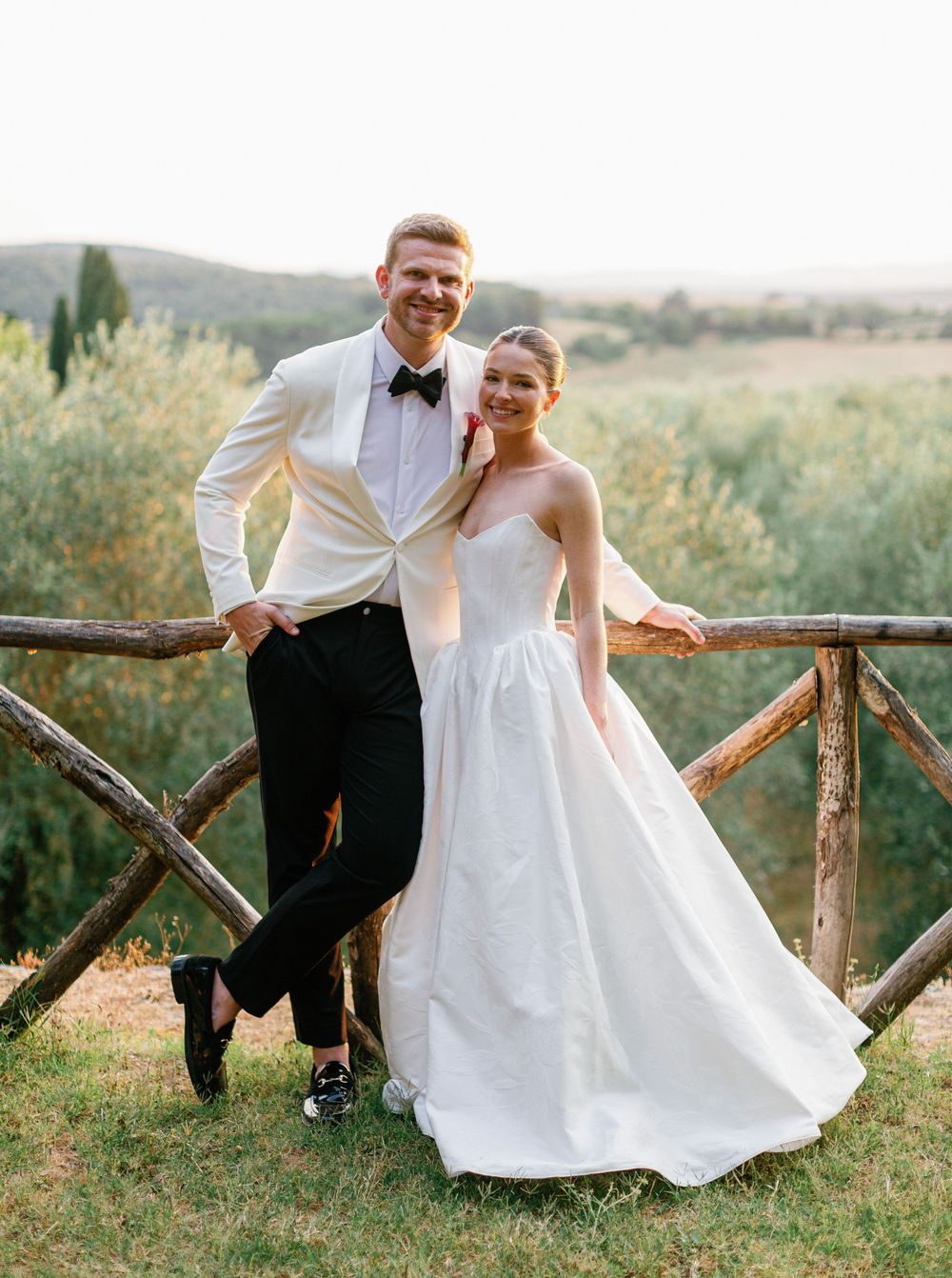 A couple in wedding attire pose playfully on wooden rustic fencing against a scenic countryside backdrop.