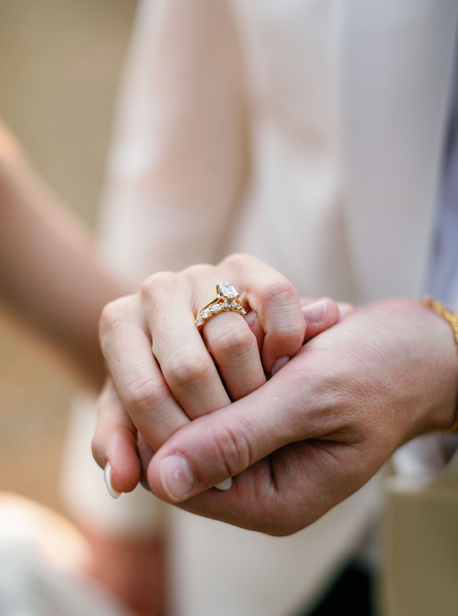Close-up view of hands showing off an engagement ring on the ring finger, set against a blurred background.