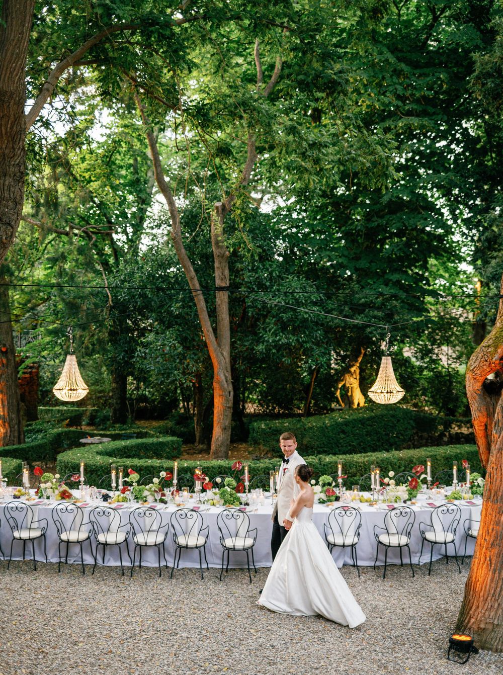 Elegant outdoor wedding reception setup in a wooded grove with hanging lights and grey chairs on gravel ground.