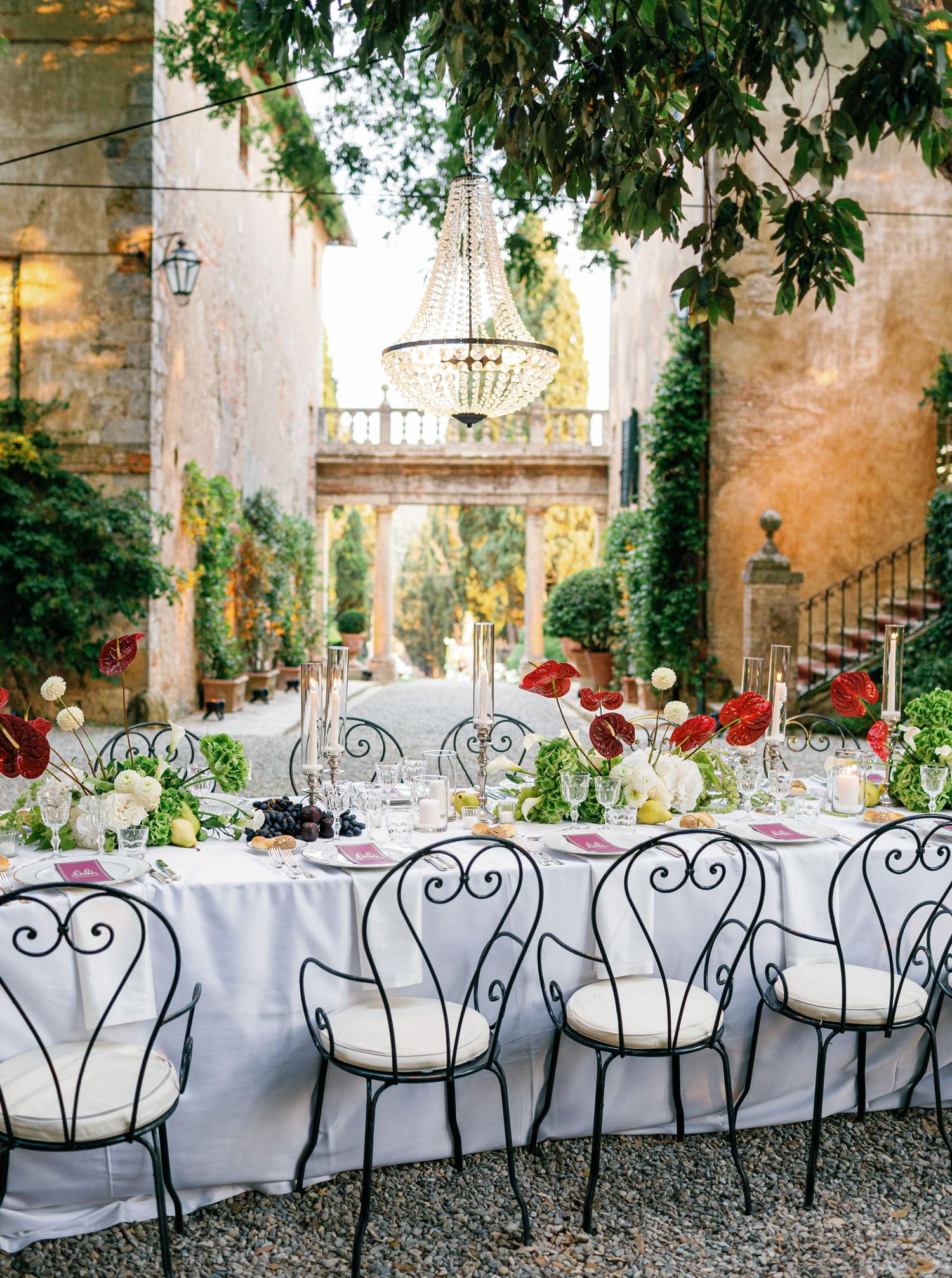 Elegant outdoor wedding reception setup at Borgo Stomennano with ornate wrought iron chairs and long white-clothed tables under trees.