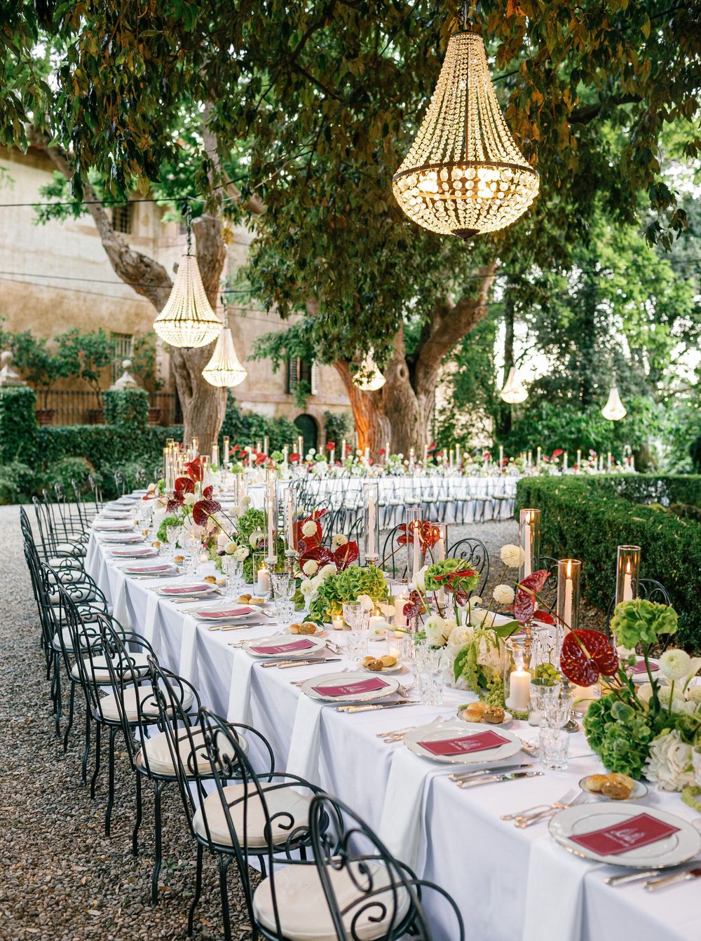Elegant long table settings with white linens and hanging rattan chandeliers under garden trees for an outdoor wedding reception in Tuscany.