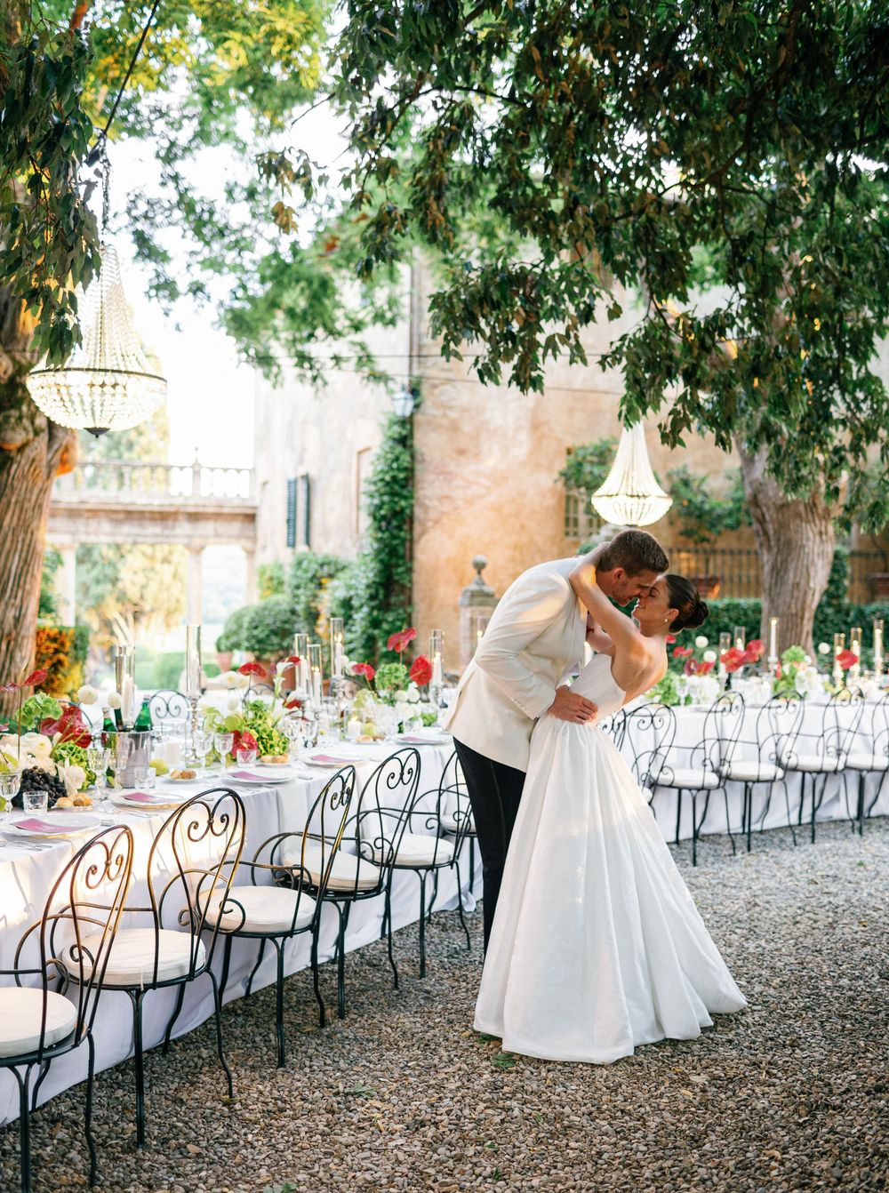 Romantic couple shares intimate wedding moments in outdoor garden setting with white tables and iron chairs under trees at Borgo Stomennano.