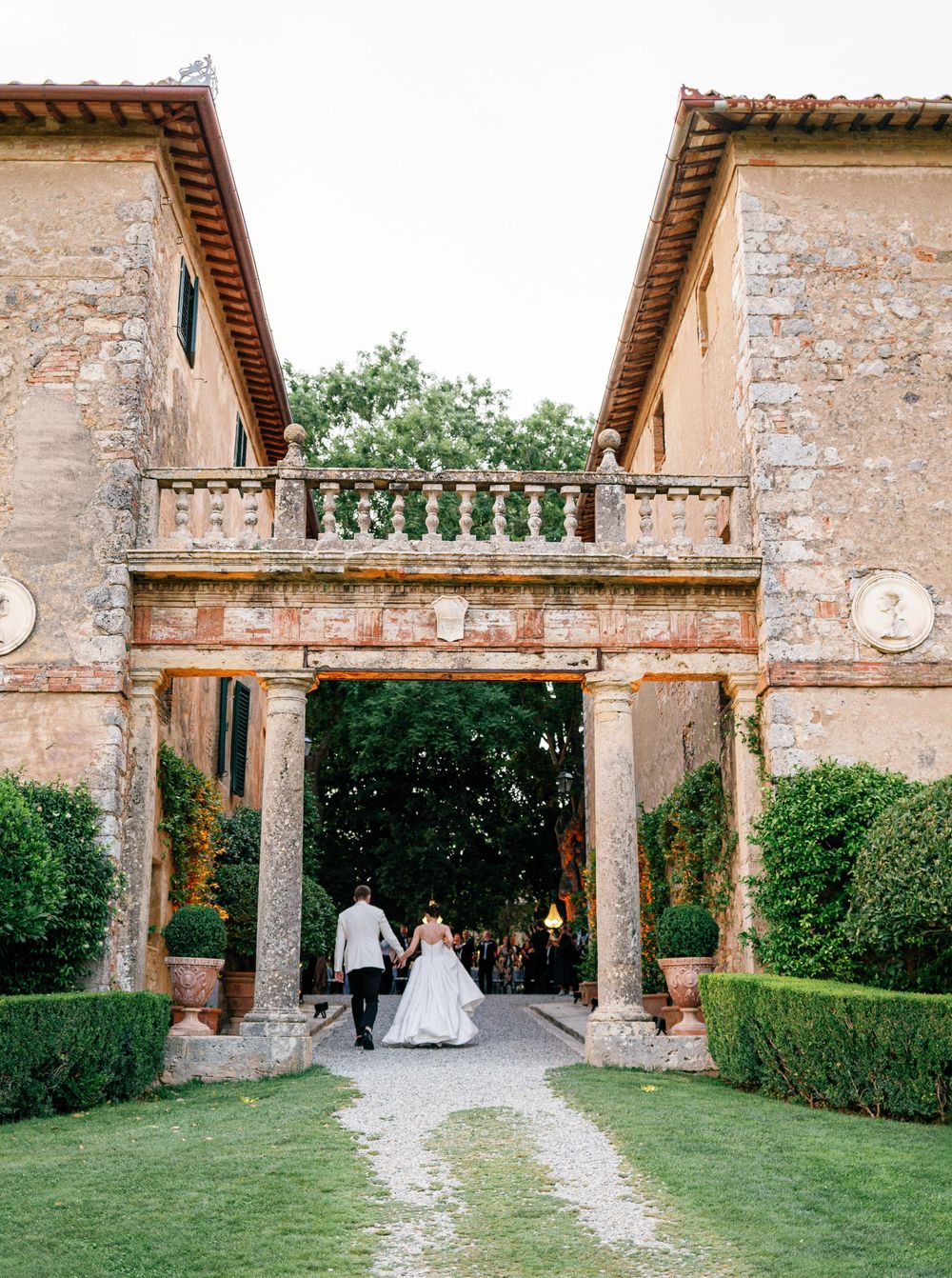 Two figures walk through a rustic Italian villa Borgo Stomennano archway with stone walls and manicured hedges on a sunny day.