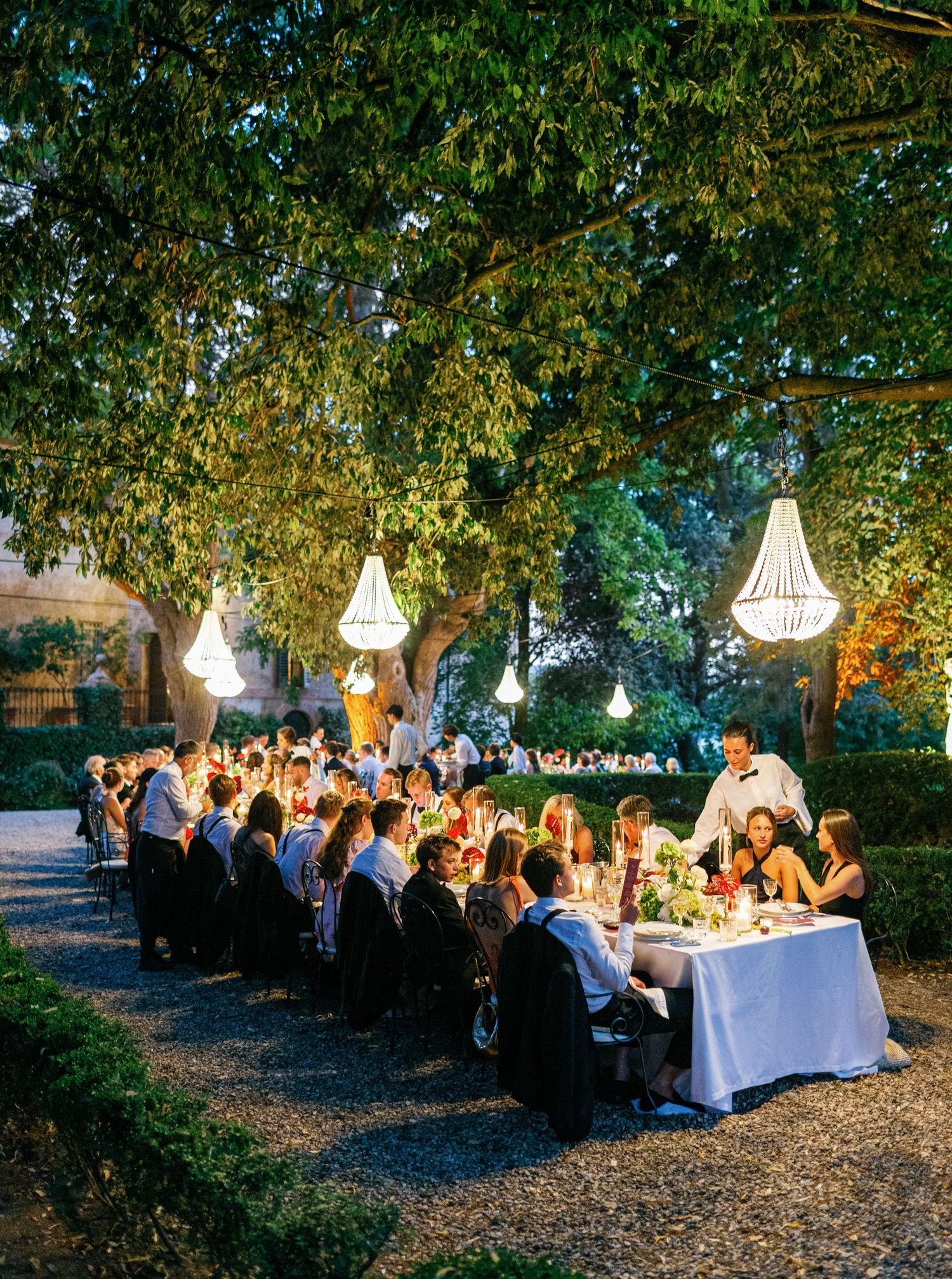 An elegant outdoor dinner party under trees  at Borgo Stomennano with suspended white lampshades and guests seated at long tables.