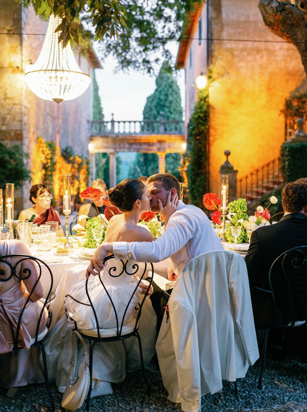 Elegant outdoor wedding reception at dusk with chandeliers and wrought iron chairs in a Borgo Stomennano villa courtyard.