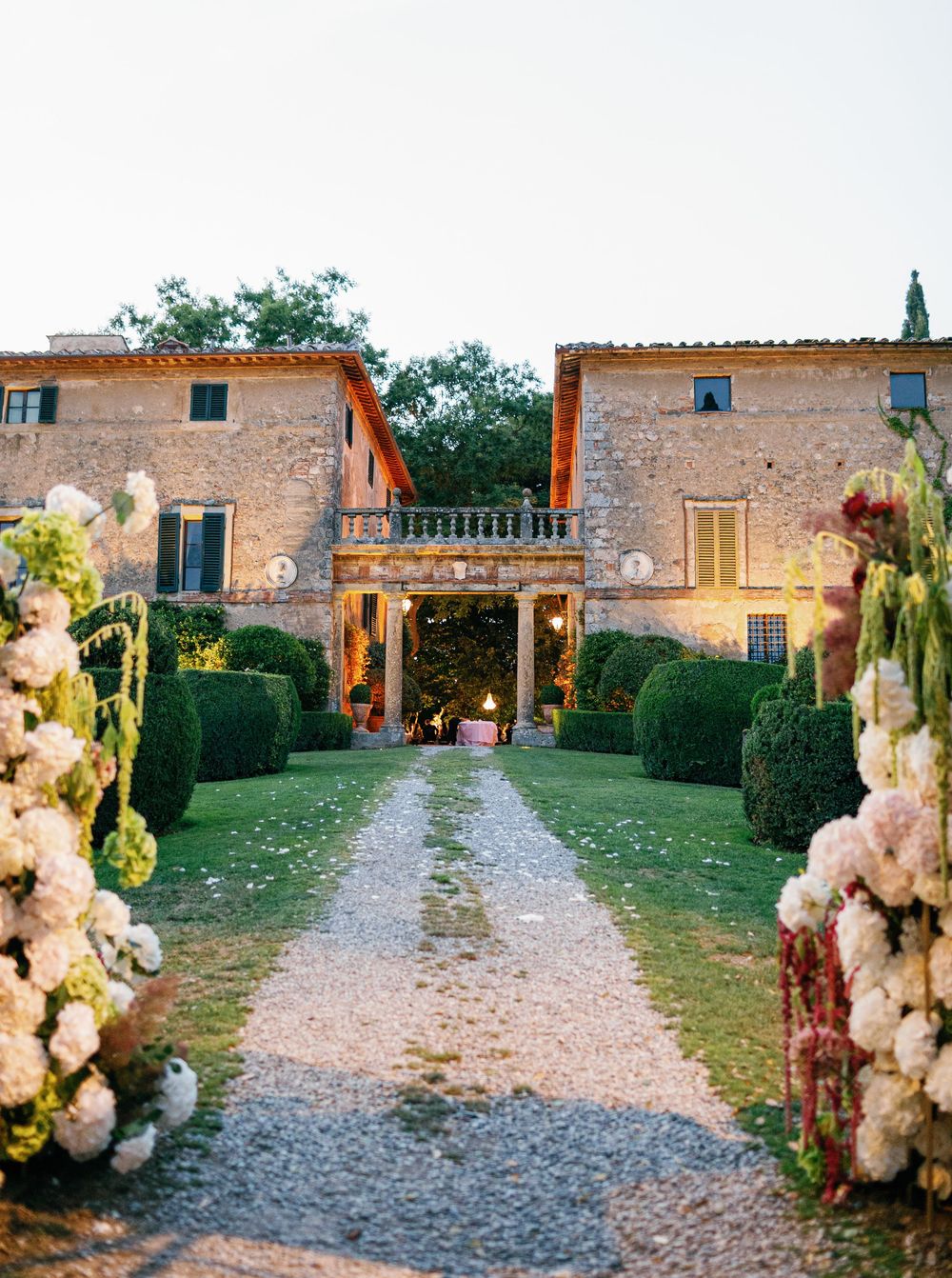 Stone-built rustic Italian courtyard at Borgo Stomennano with manicured hedges and cream-colored flowers along a gravel pathway at sunset.