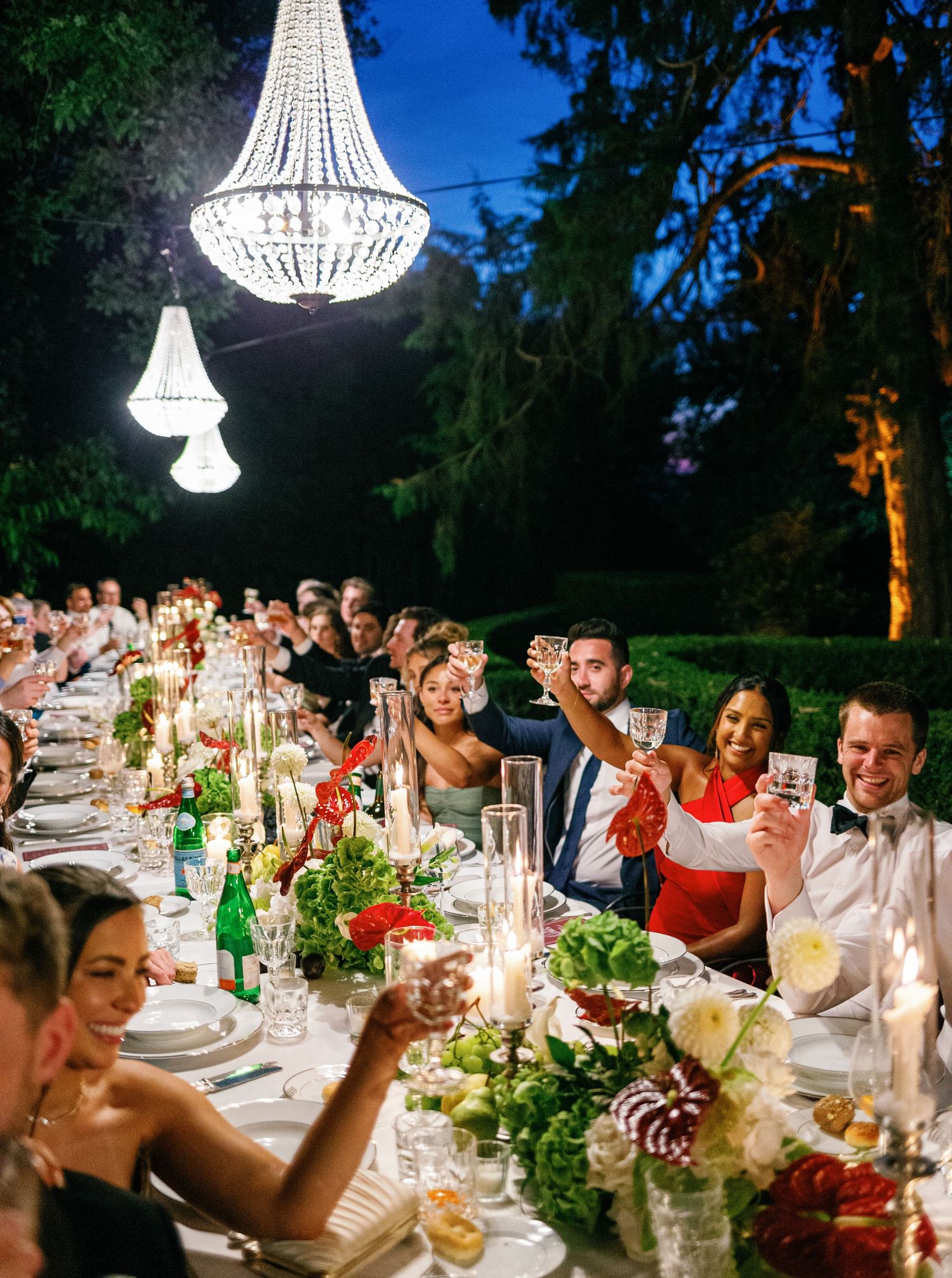 Elegant outdoor dinner party with crystal chandeliers illuminating long tables adorned with floral arrangements at dusk.