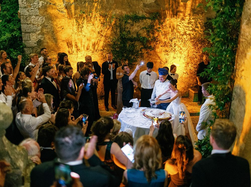 A crowd of wedding guests celebrate outdoors at night with illuminated stone walls creating a warm ambiance.