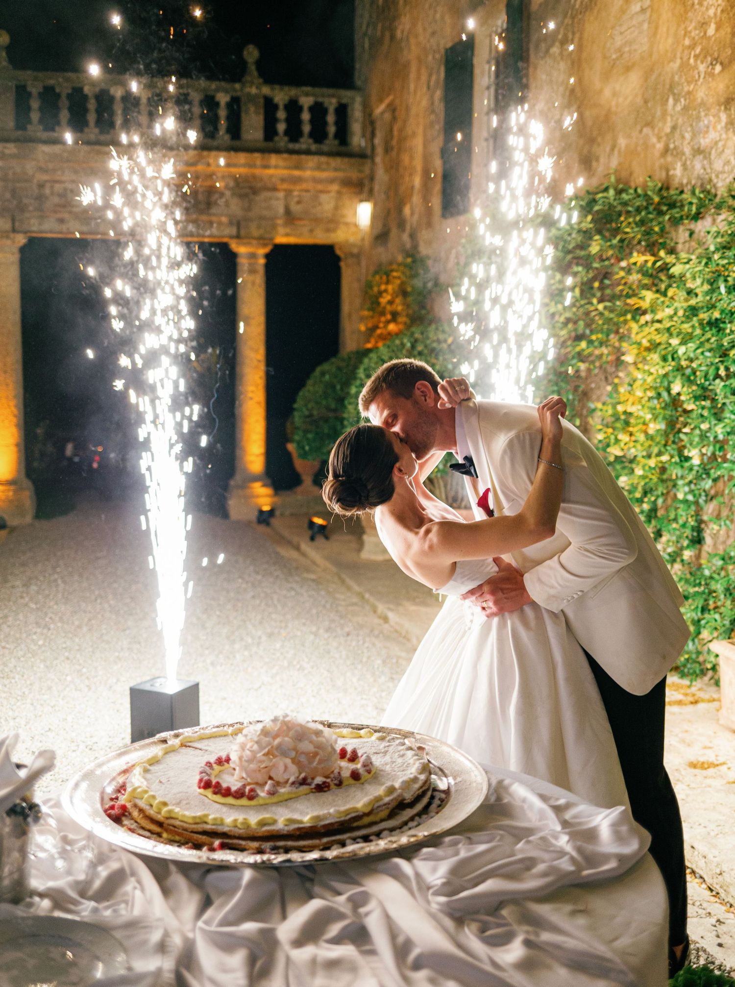 A romantic evening wedding cake cutting scene with sparklers illuminating an outdoor courtyard venue.