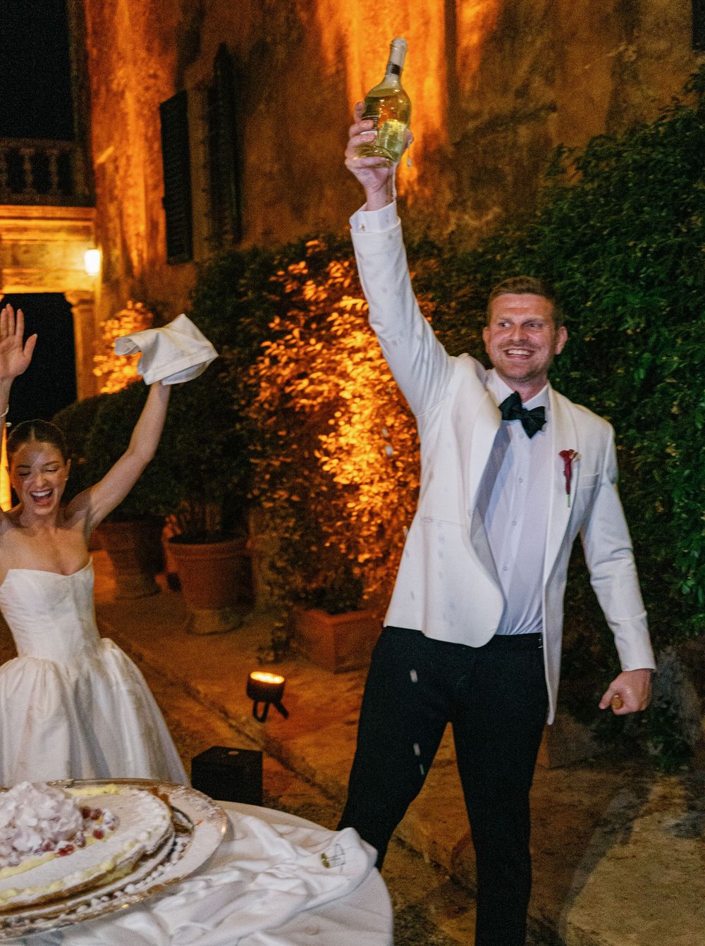 Joyful couple celebrates at a wedding reception with raised champagne glasses in a warmly lit rustic courtyard setting.