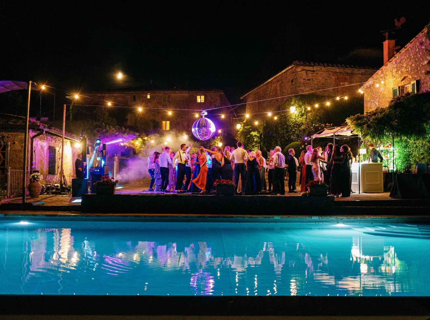 String lights illuminate a romantic outdoor evening gathering with a glowing blue swimming pool and nighttime ambiance.
