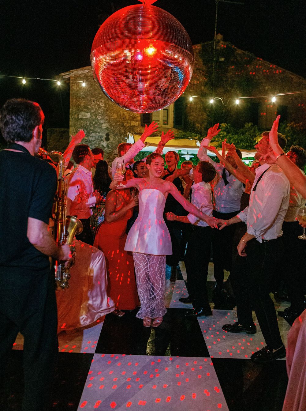 Wedding party dance beneath a glowing red disco ball at a nighttime outdoor party with string lights overhead.