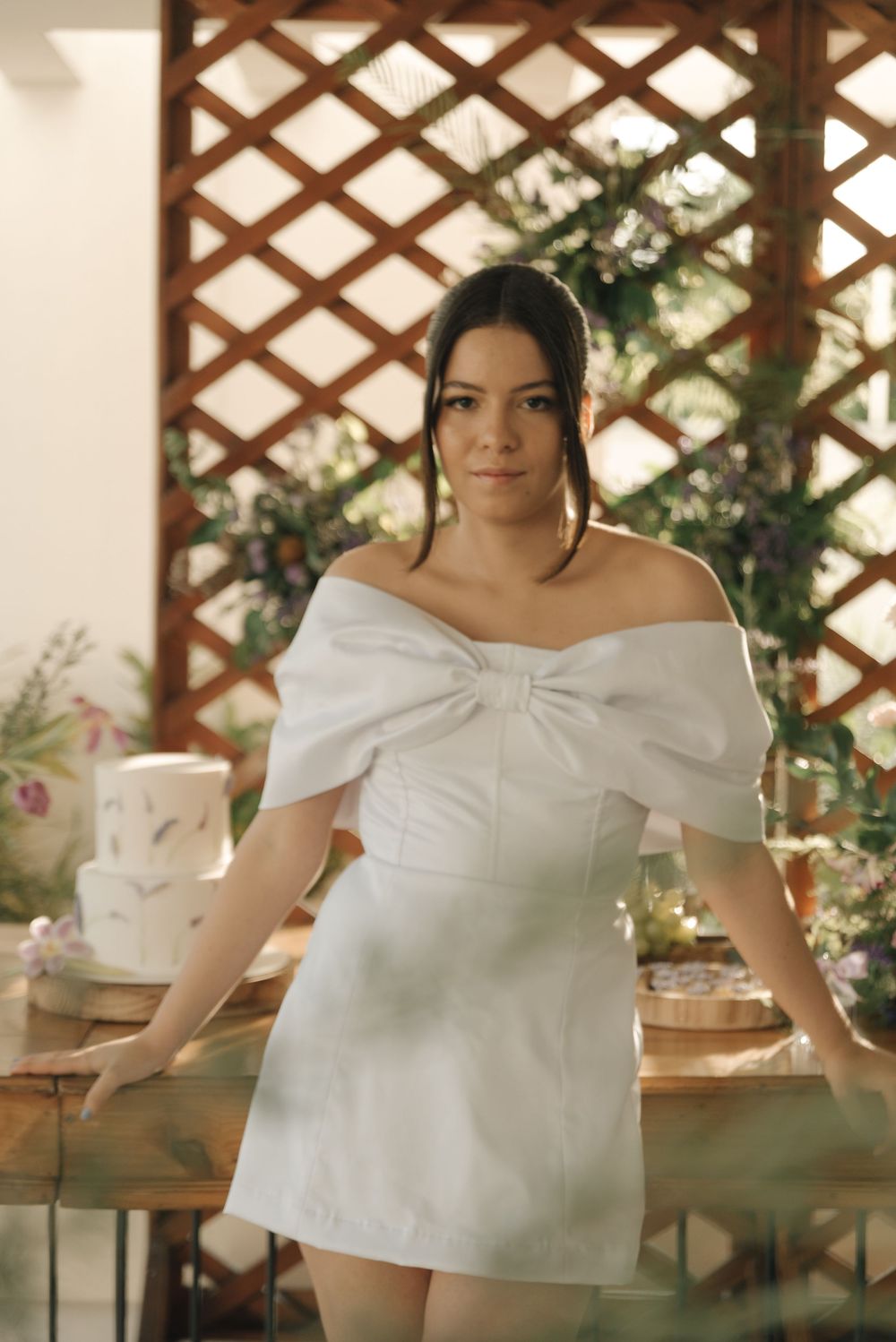A series of photos showing a white off-shoulder top with bow detail against a wooden lattice background with greenery.